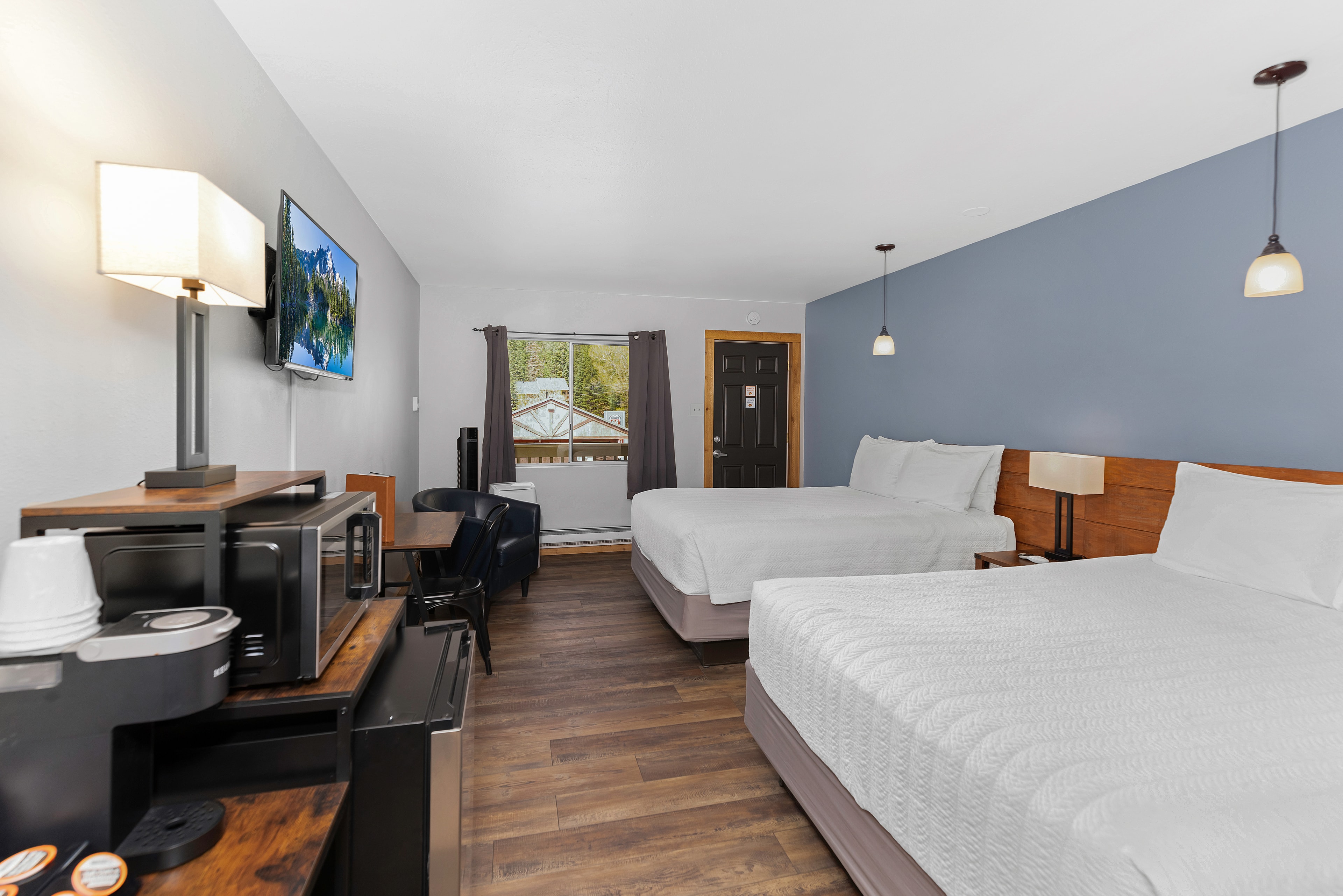 A view from the corner of the room looking toward the entry door and window. The foreground features a coffee maker and microwave on a wooden stand, while the two queen beds occupy the right side of the room under soft pendant lighting.