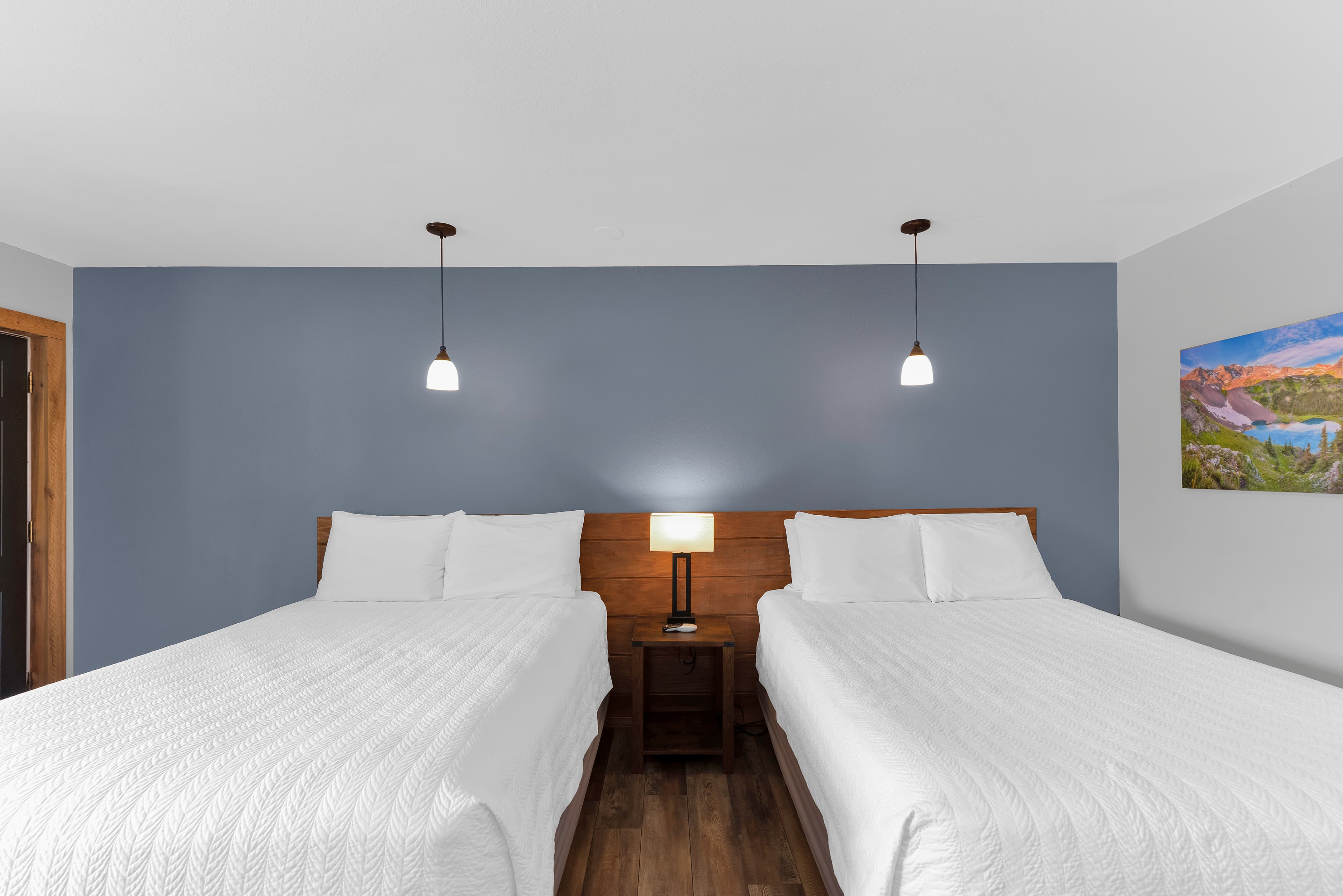 A symmetrical front view of two queen beds with white textured coverlets and a shared wooden headboard. The beds are set against a dusty blue accent wall with two hanging pendant lights and a central nightstand lamp.