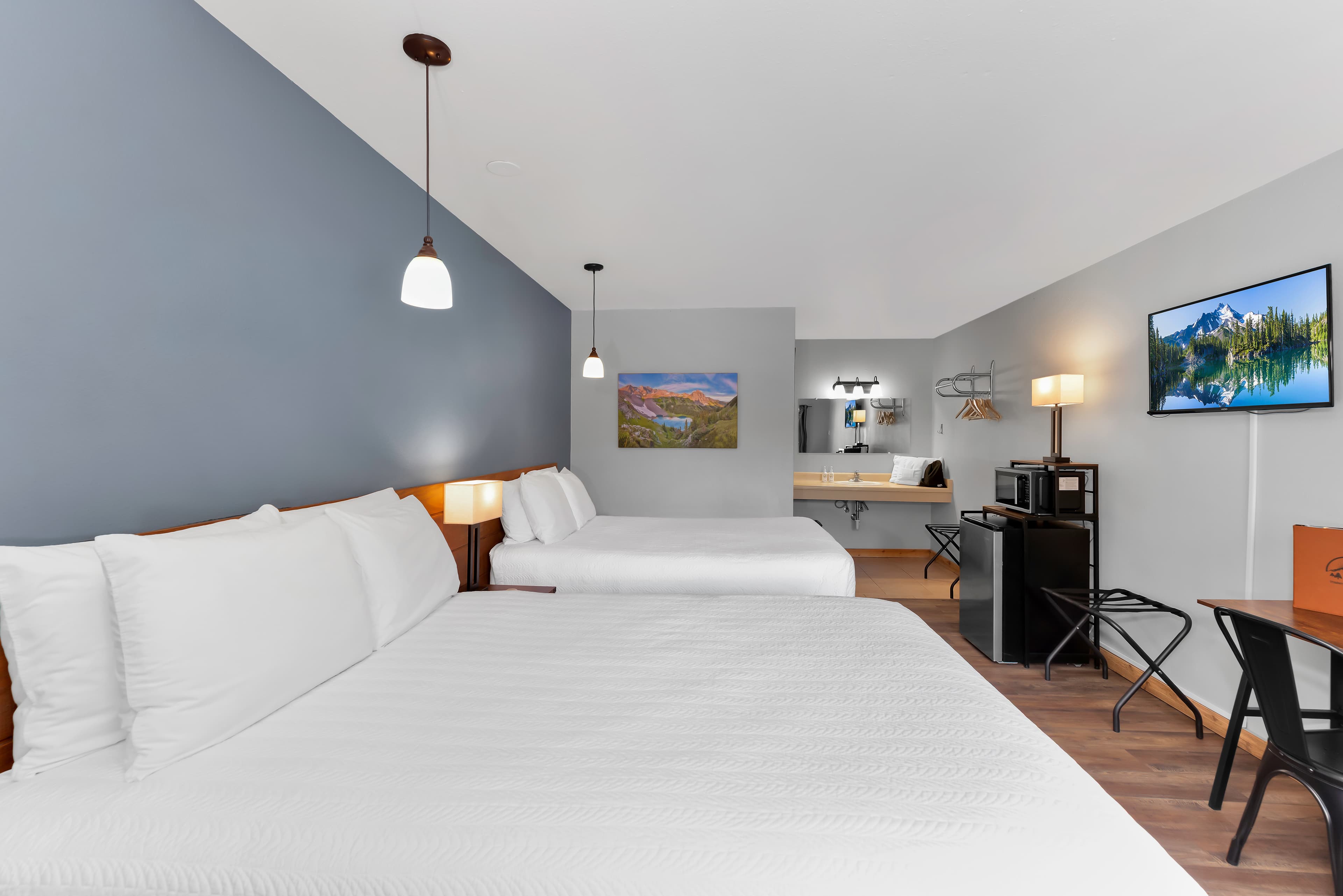 A side-angle view of the guest room showing the two queen beds and the functional living space. Along the right wall, there is a flat-screen TV, a luggage rack, and a small kitchenette station with a mini-fridge and microwave, leading toward an open vanity area in the back.