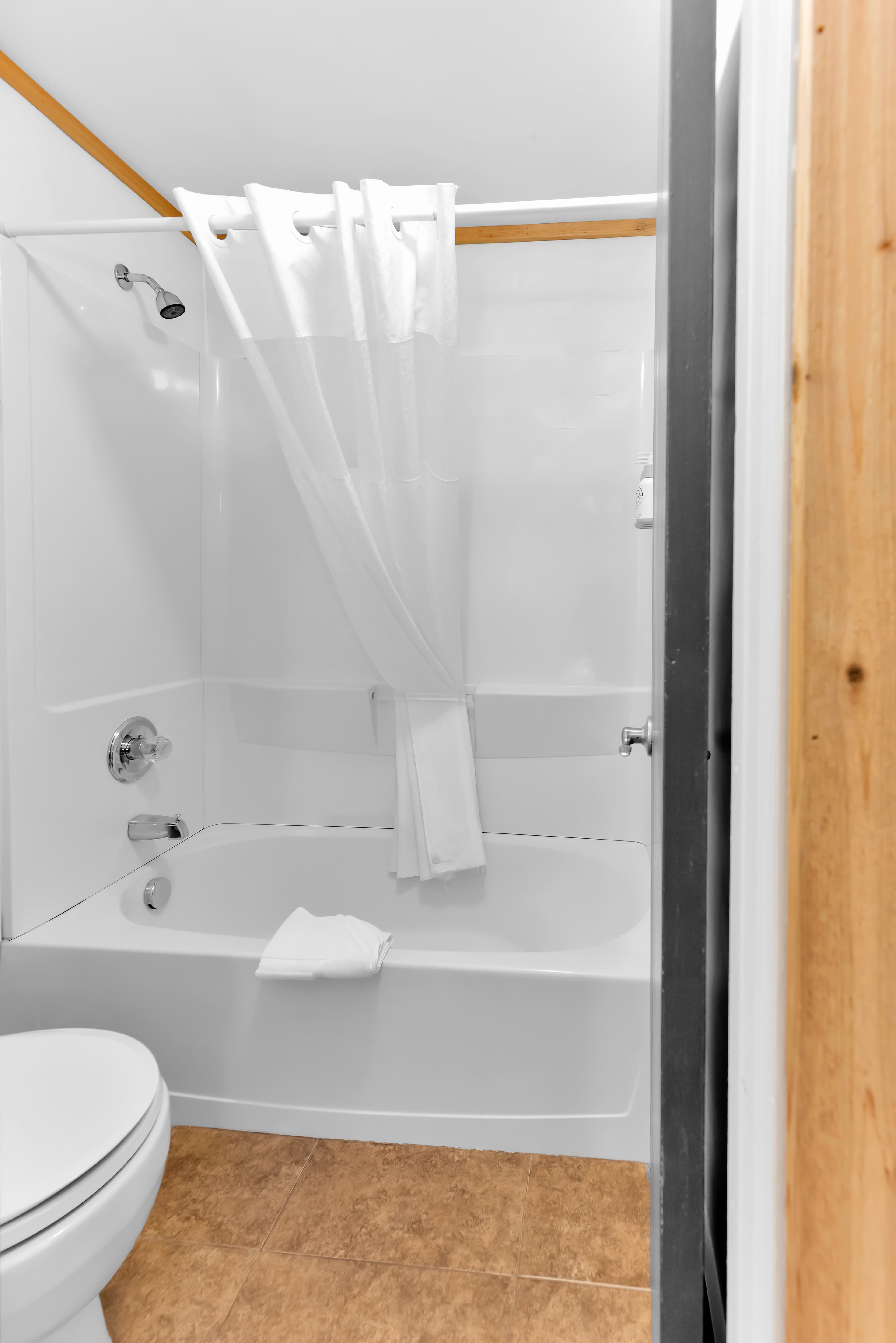 A bright bathroom featuring a white alcove bathtub and shower with a white curtain. The space includes tan floor tiles and a partial view of a white toilet, framed by a wooden door jam.