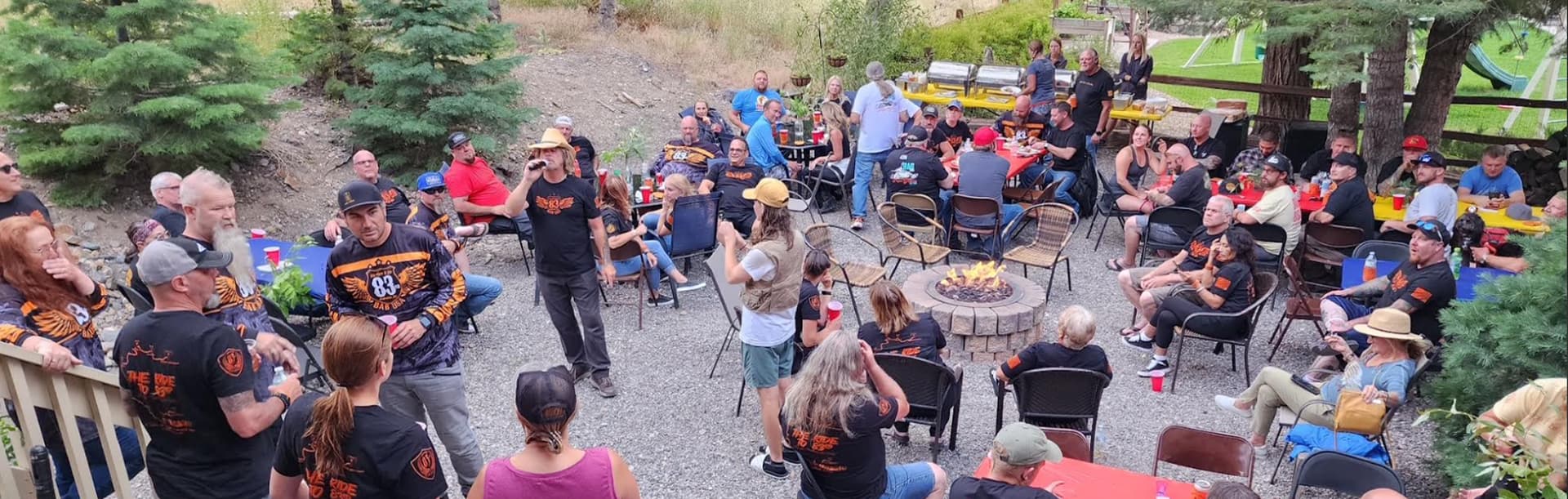 A lively outdoor gathering with a large group of people socializing around tables and a fire pit, surrounded by trees.
