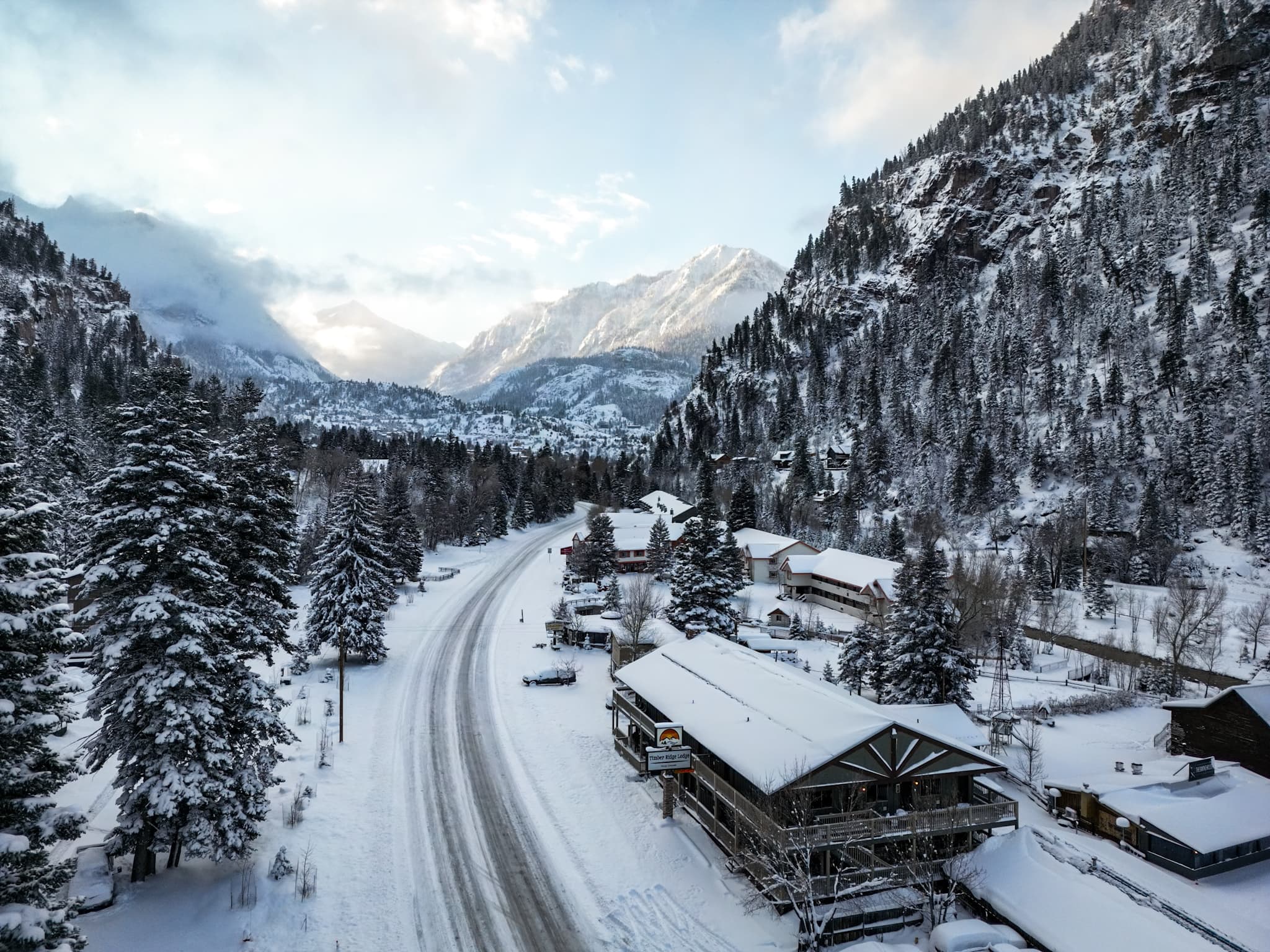 Snow-covered road winding through a mountain valley with rustic buildings and evergreen trees.