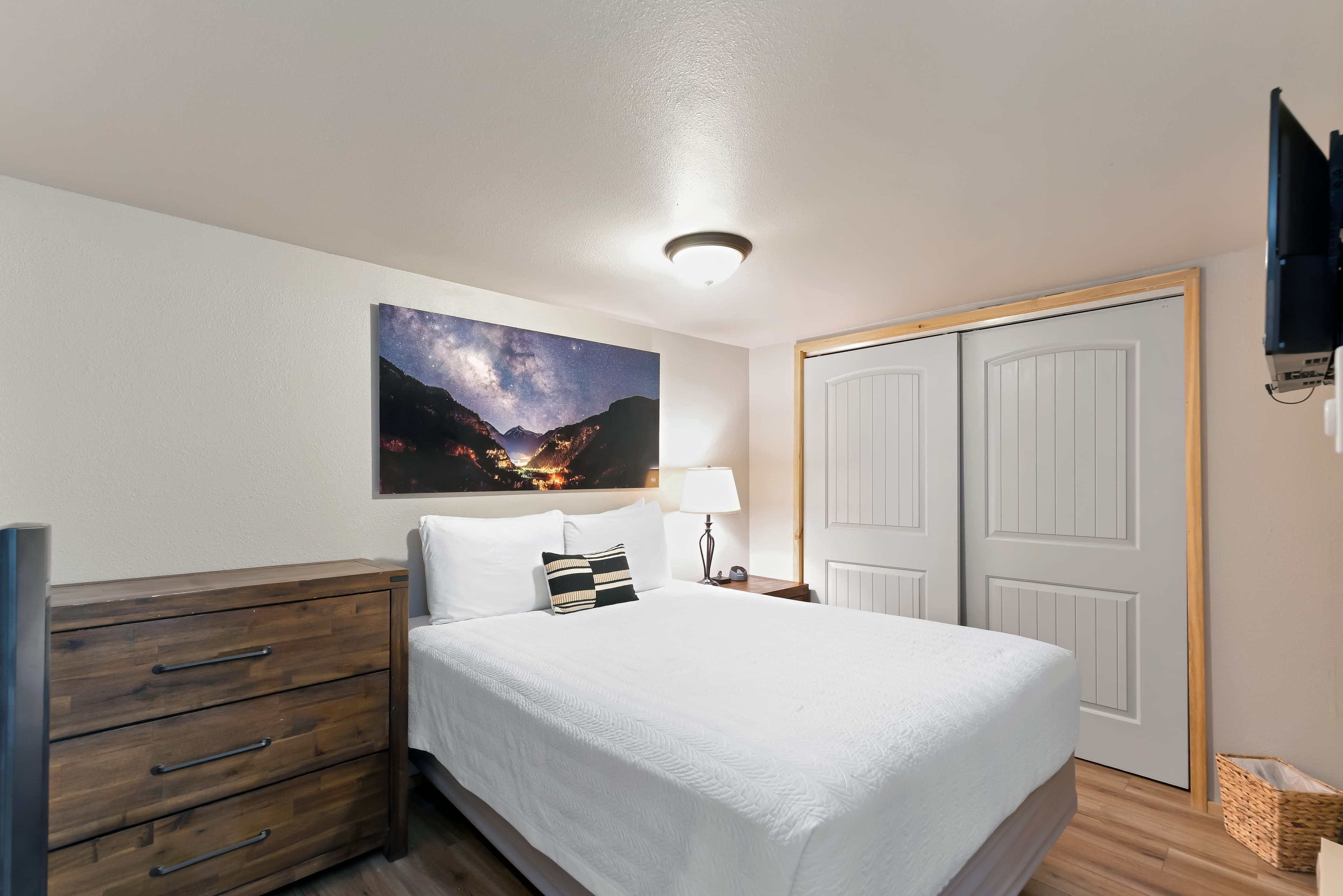 The second bedroom showing a queen bed against a grey wall with a small nightstand and lamp. The room includes a built-in nook with a chest of drawers and a framed photo of a hiker, with an open doorway leading back to the bright living area.