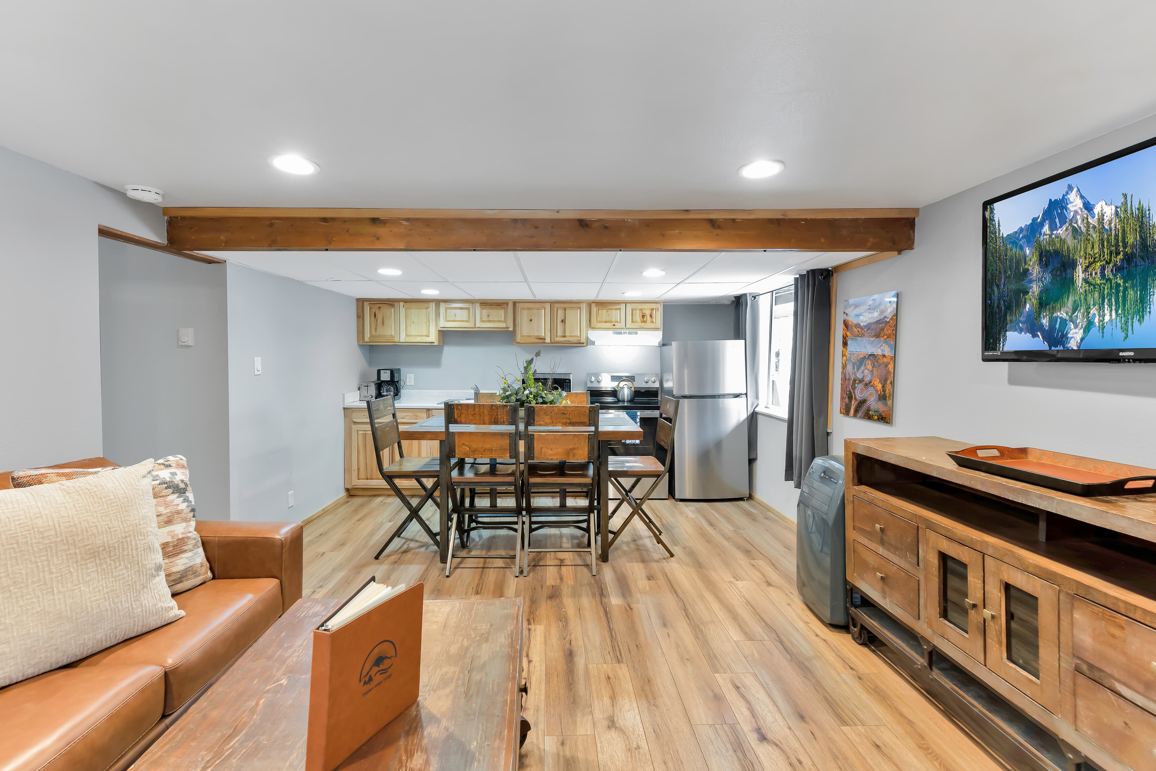 A wide view of the apartment's common area. It features a large wooden media console with a wall-mounted flat-screen TV on the right, a central dining area, and a kitchen equipped with stainless steel appliances, including a full-sized refrigerator and stove.