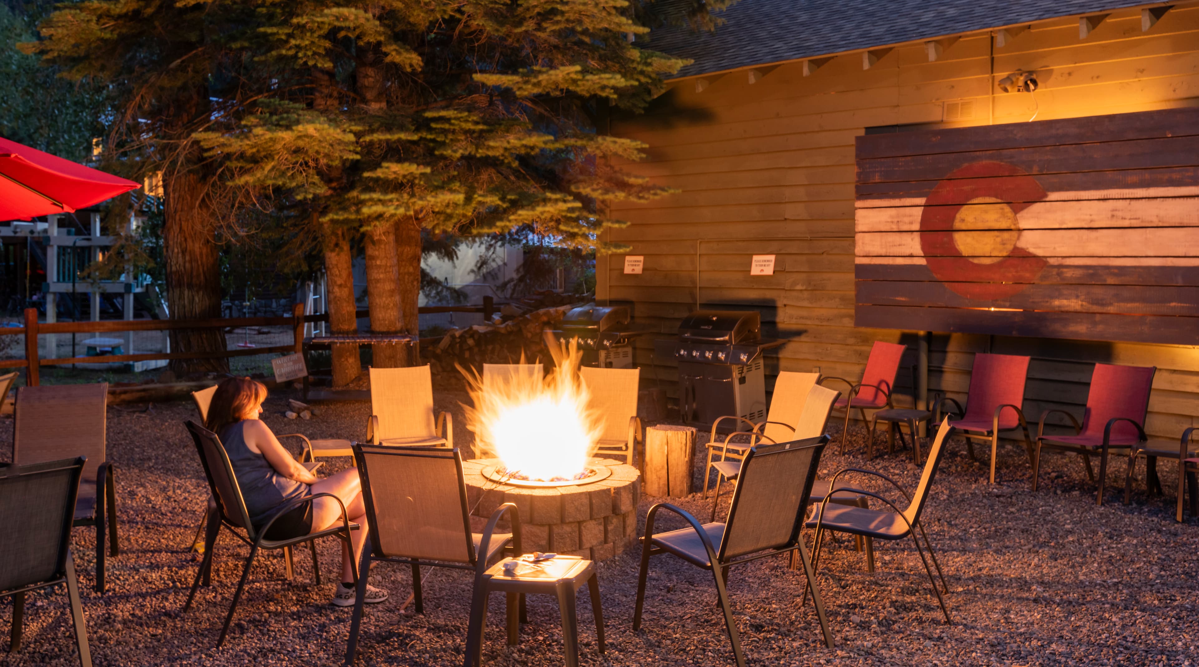 A woman sits by a fire pit surrounded by chairs in a cozy outdoor setting decorated with a Colorado flag.