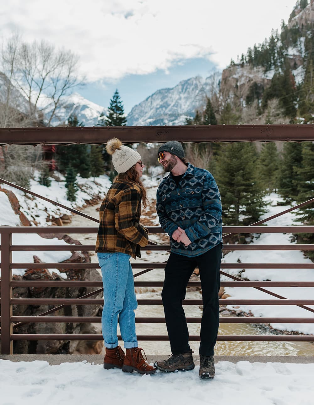 A couple playfully leans in for a kiss on a snowy bridge with mountains in the background.