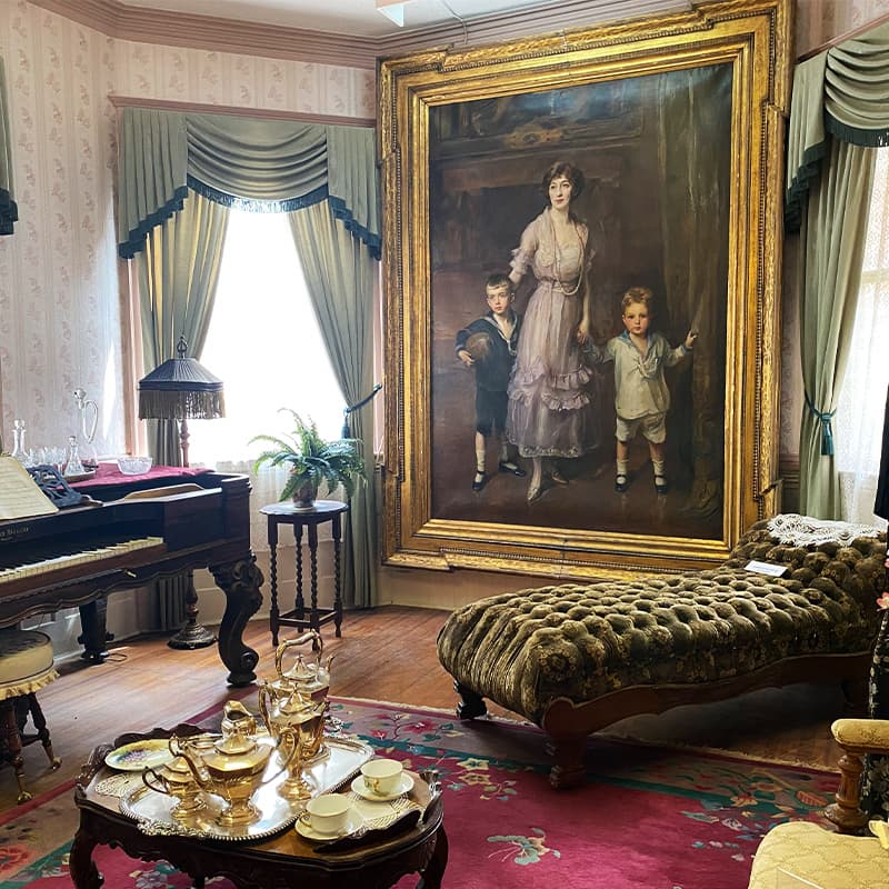 A vintage living room featuring a large portrait of a woman with two children, alongside a piano and ornate furniture.