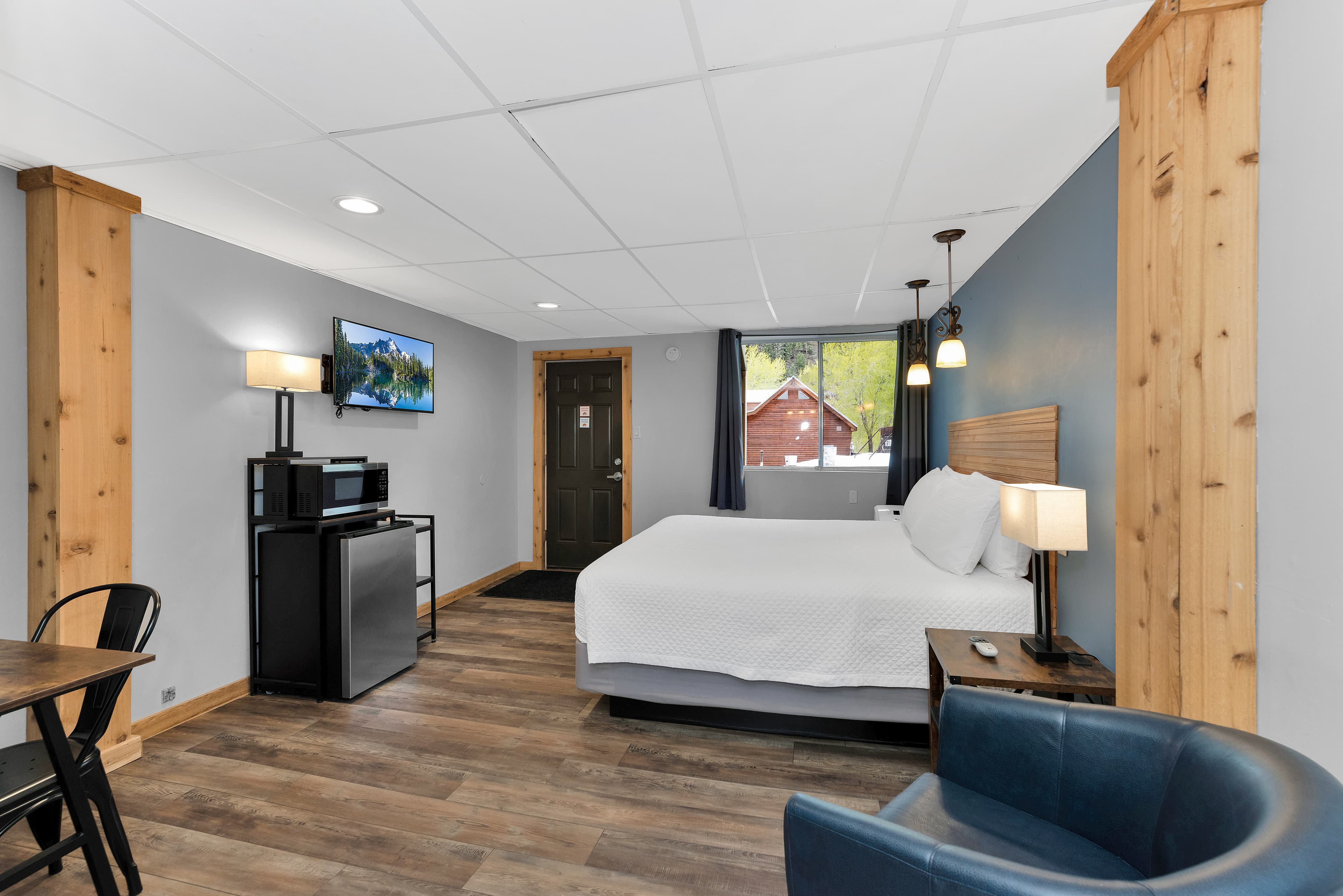 A wide view of the King Room featuring a king-sized bed with white linens and a wooden headboard. The room includes wood-look flooring, a blue leather armchair in the foreground, and a kitchenette station with a microwave and mini-fridge beneath a wall-mounted TV.