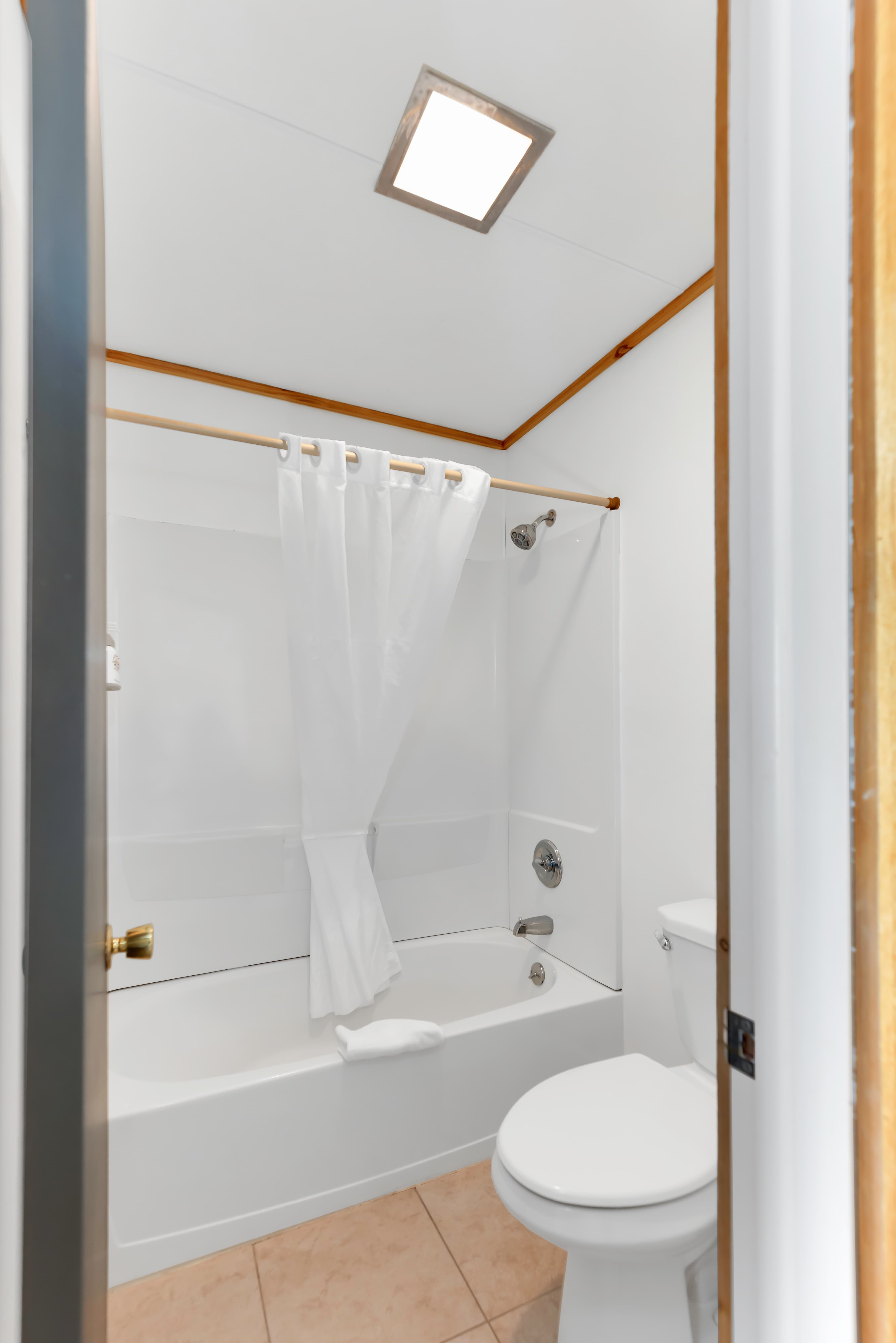 A clean, bright bathroom featuring a white bathtub and shower combination with a white curtain. The room includes a standard white toilet, tan floor tiles, and a skylight providing natural illumination.