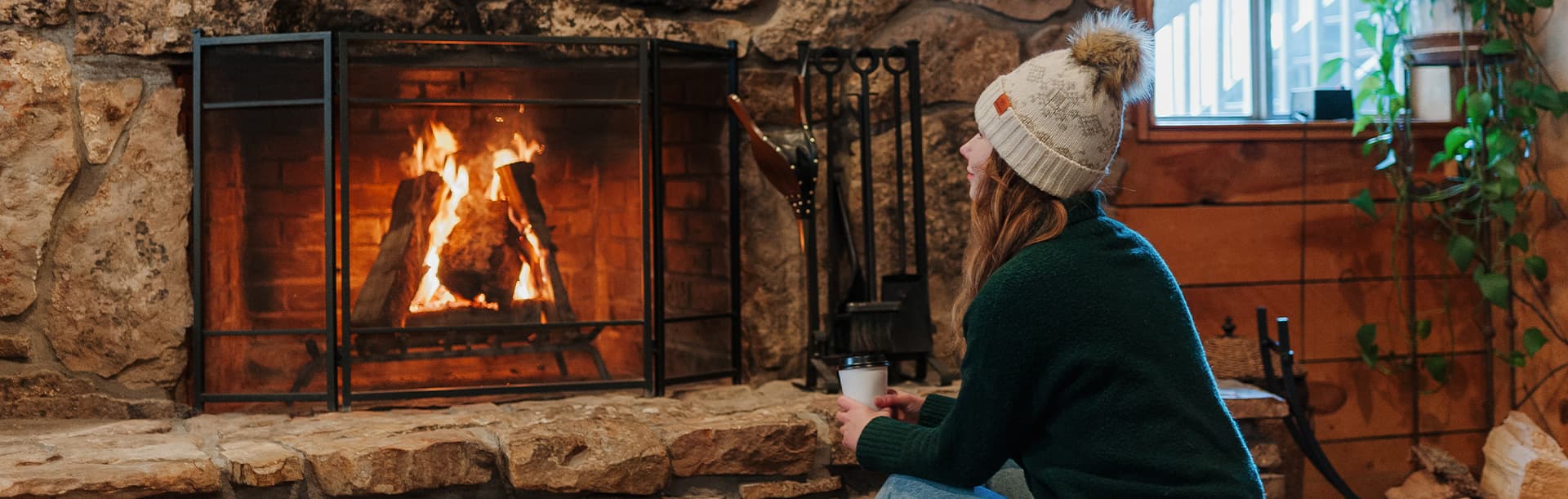 A person sits by a cozy fireplace, holding a cup and wearing a winter hat.