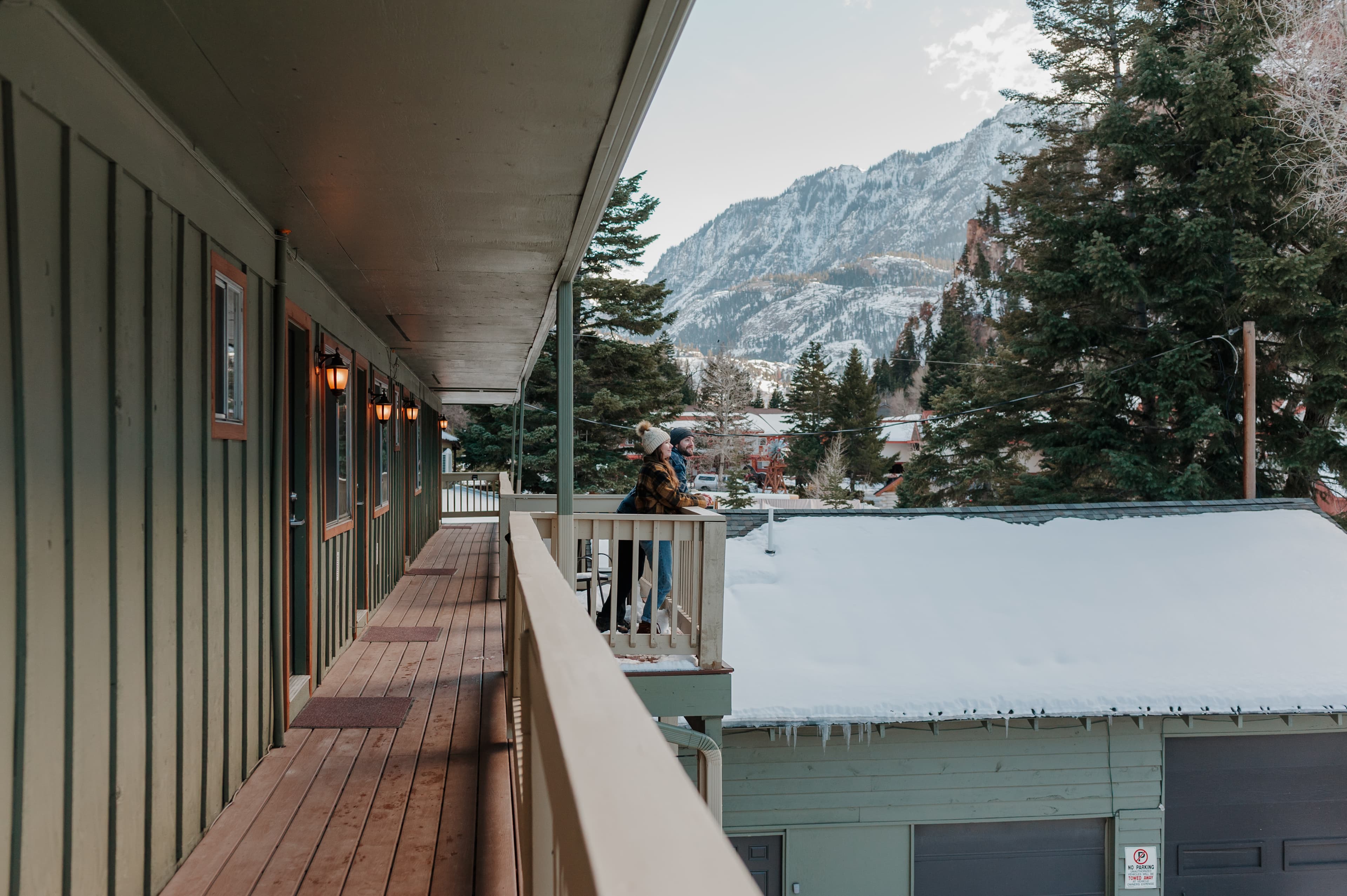 A person stands on a balcony overlooking snow-covered rooftops and mountains in the background.