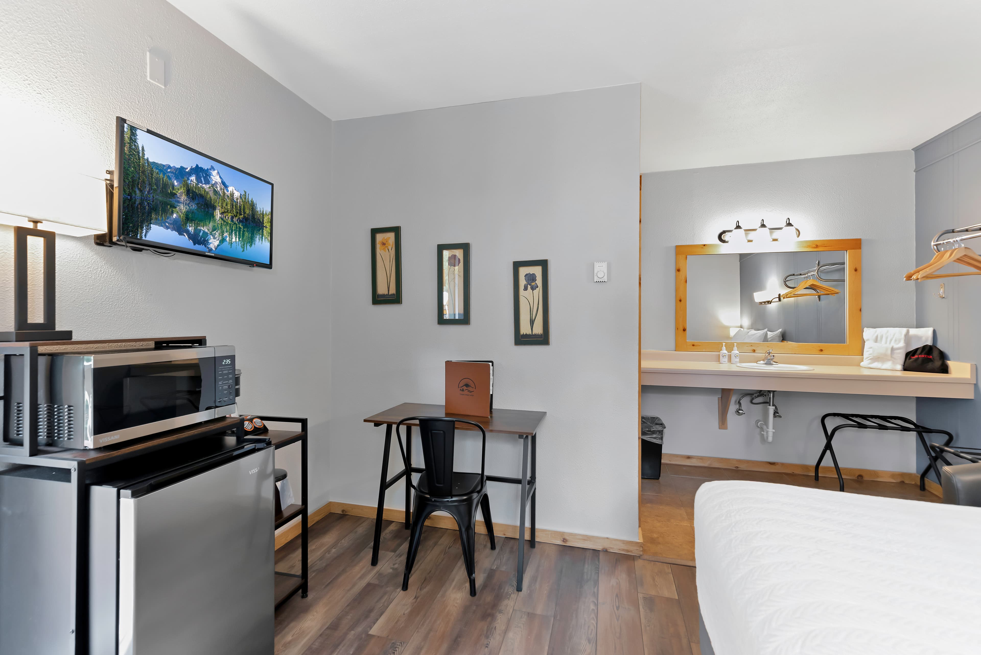 A functional side view of the Queen Room highlighting essential travel amenities including a wall-mounted flat-screen TV, a mini-fridge and microwave station, and a small writing desk. The open-concept vanity area features a large mirror and natural wood accents.