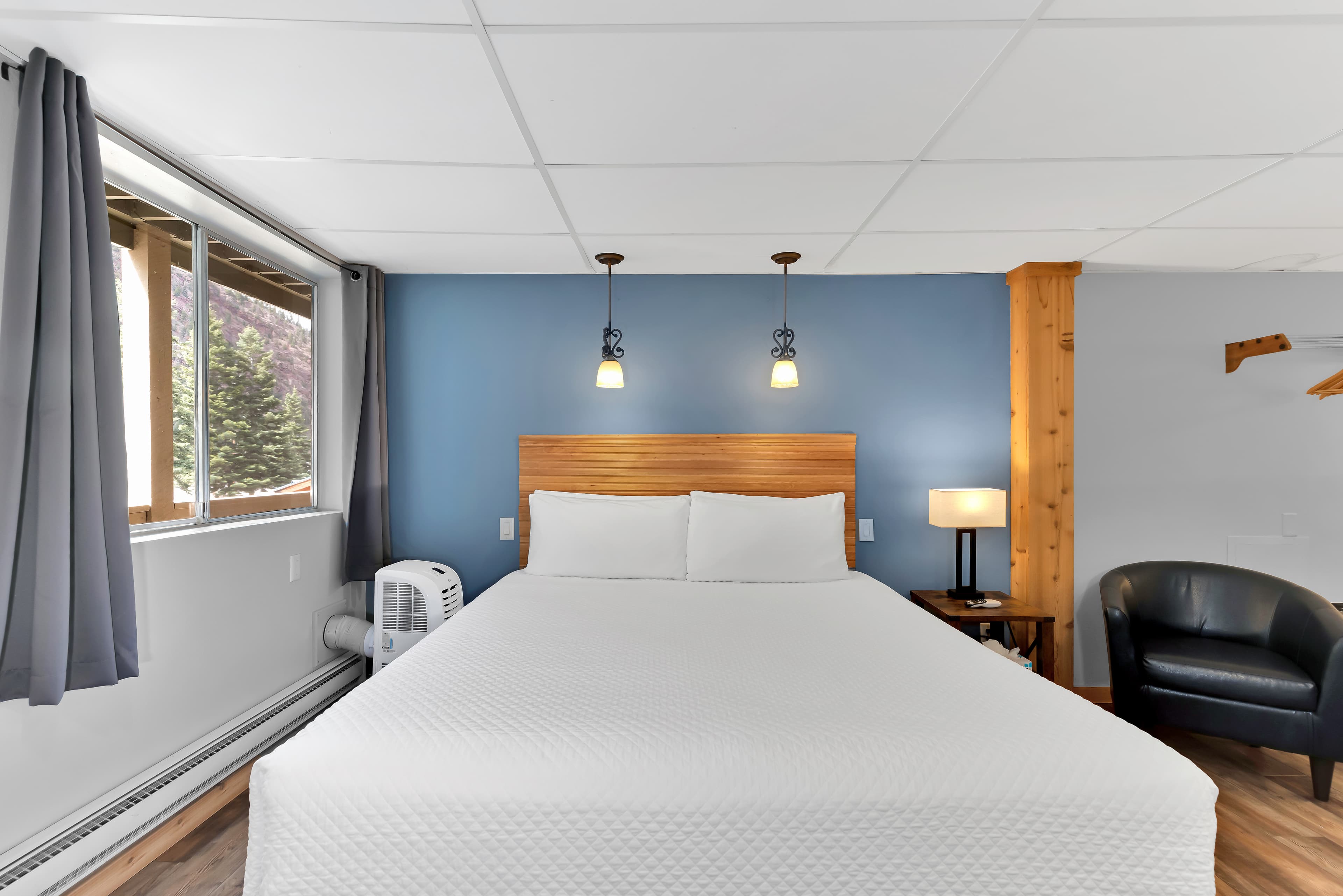 A straight-on view of the king bed centered against a blue accent wall with two hanging pendant lights. To the left is a large window with grey curtains and a portable AC unit; to the right is a rustic wood pillar and a black leather barrel chair.
