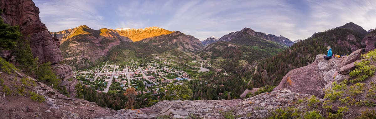 A panoramic view of a mountain town surrounded by lush greenery and towering peaks at sunset.