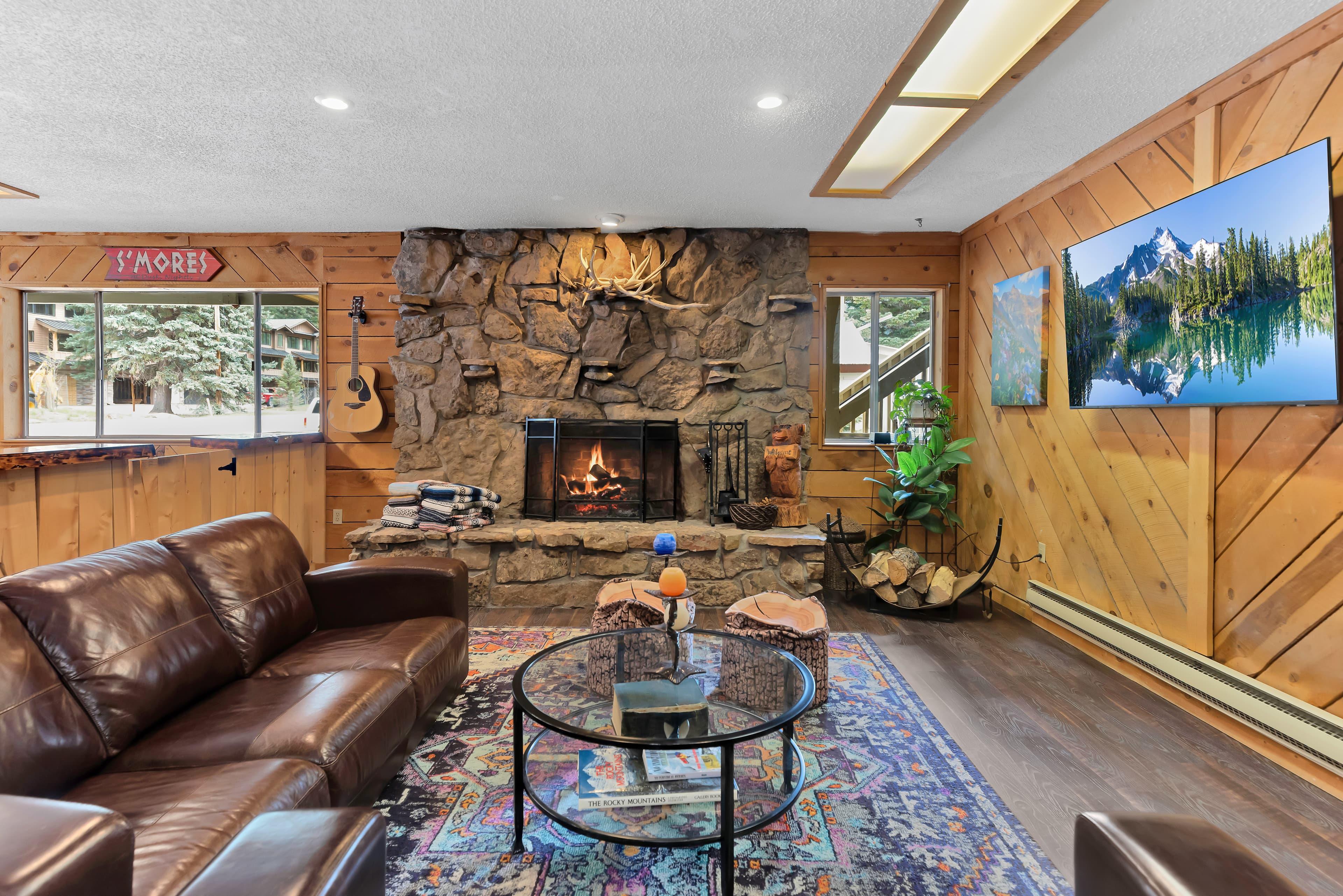 A cozy living room featuring a stone fireplace, leather sofa, and a mounted TV, surrounded by wooden walls and large windows.