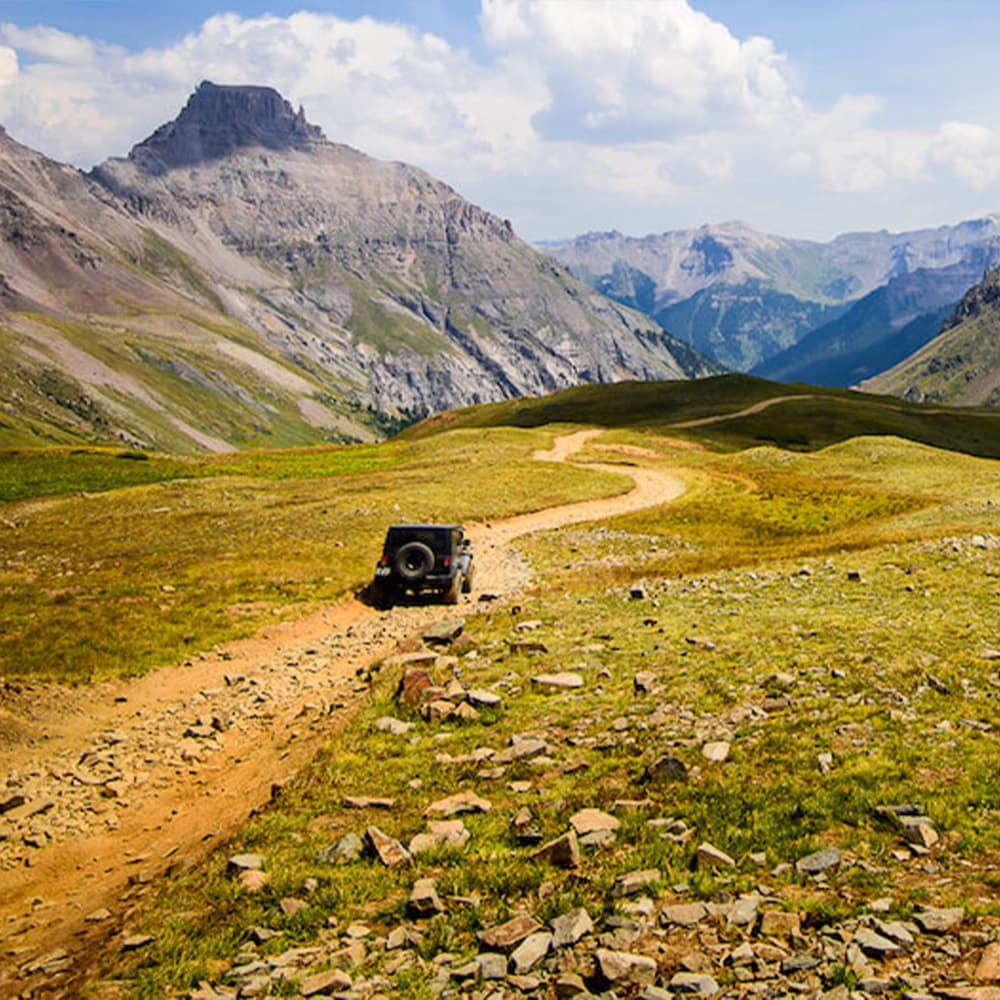 A jeep drives along a winding dirt path in a mountainous landscape.