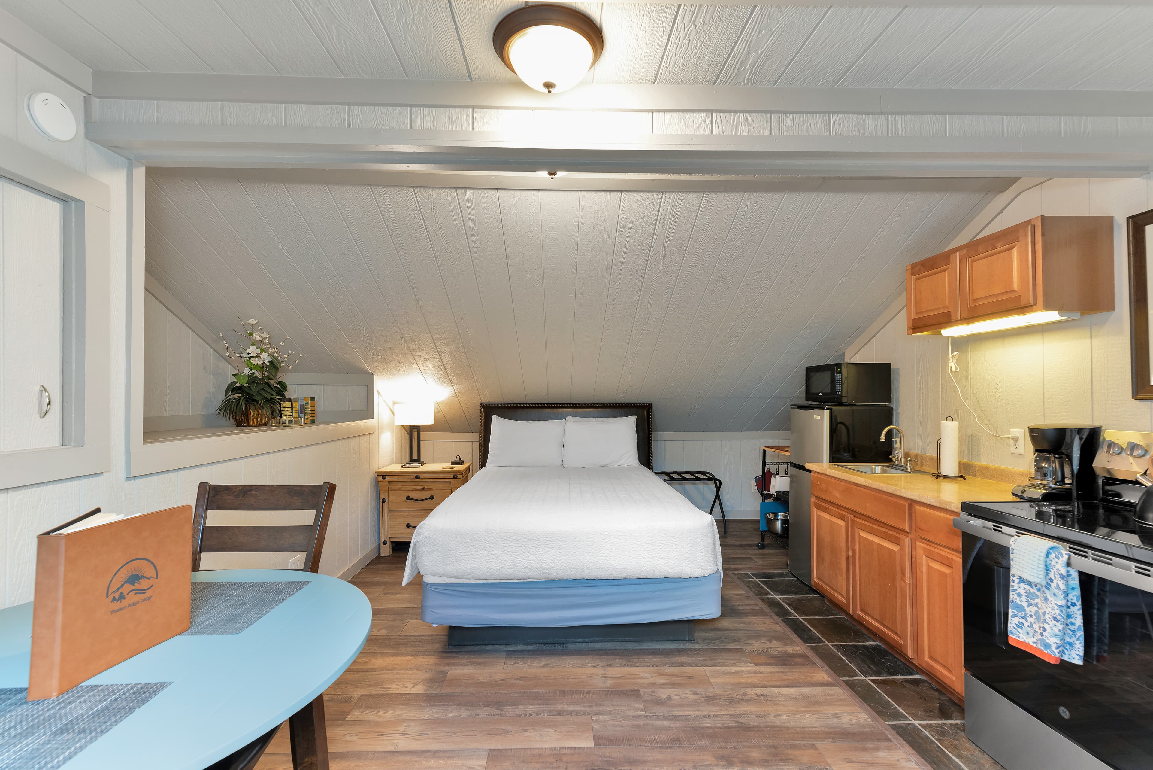 Interior of a cozy mountain loft suite at Timber Ridge Lodge in Ouray, CO. The room features a queen bed tucked under a vaulted, white-paneled ceiling, a full kitchenette with wooden cabinets and a stove, and a small blue dining table on wood-style flooring.
