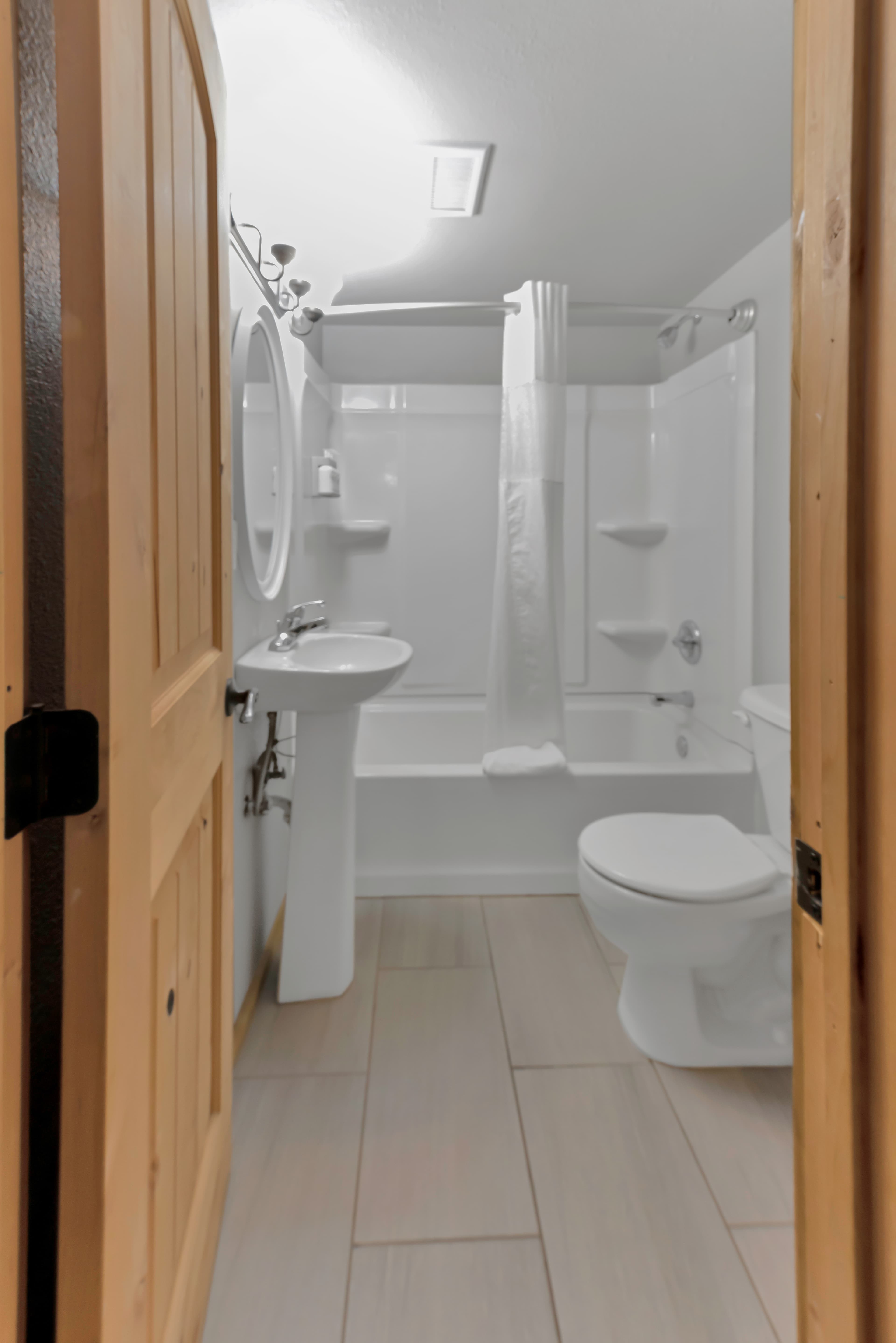 A view through a wooden door into a clean bathroom. It includes a white pedestal sink, a large oval mirror, a toilet, and a bathtub with a white shower curtain. The floor is finished with large tan rectangular tiles.