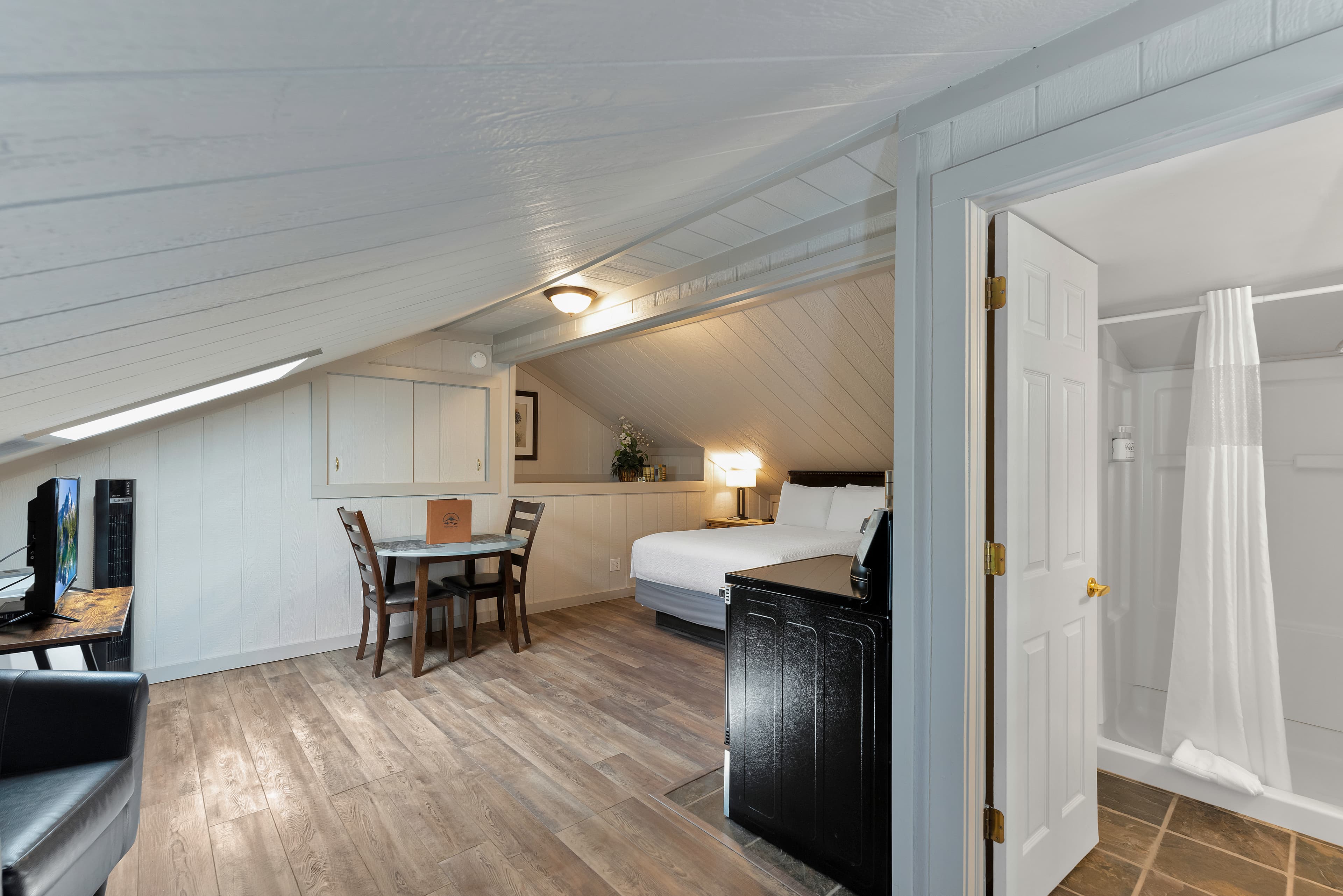 A wide-angle view of the Timber Ridge Lodge loft layout showing the transition from the living area to the bathroom. The shot captures a wall-mounted TV, a dining nook, and a queen bed, with an open door revealing a clean bathroom with slate-colored tile flooring.