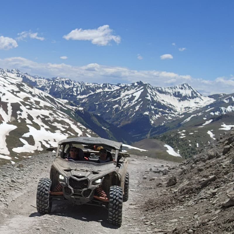 An off-road vehicle drives along a rocky mountain trail with snow-capped peaks in the background.