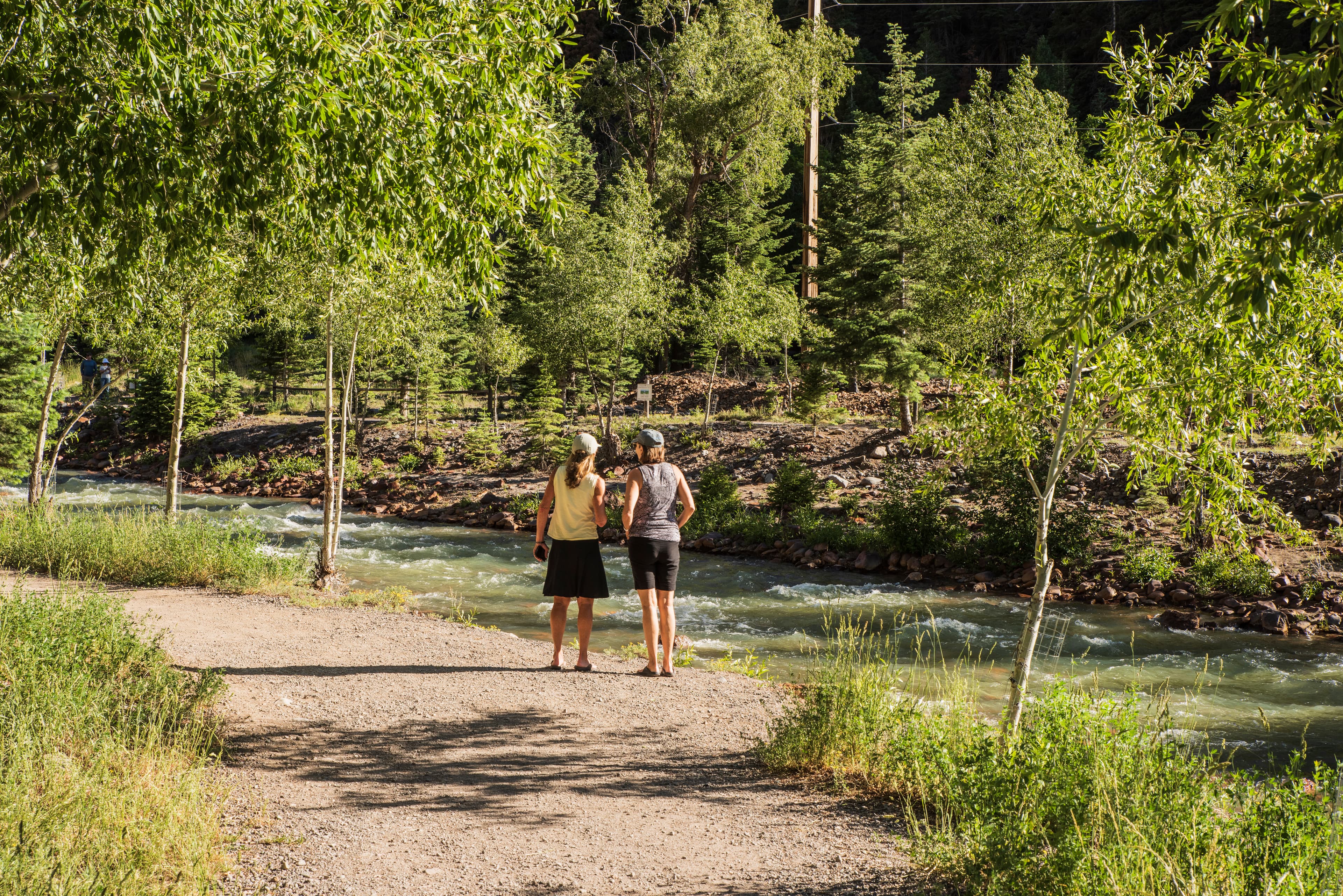 Two people stand by a river, surrounded by trees and greenery.