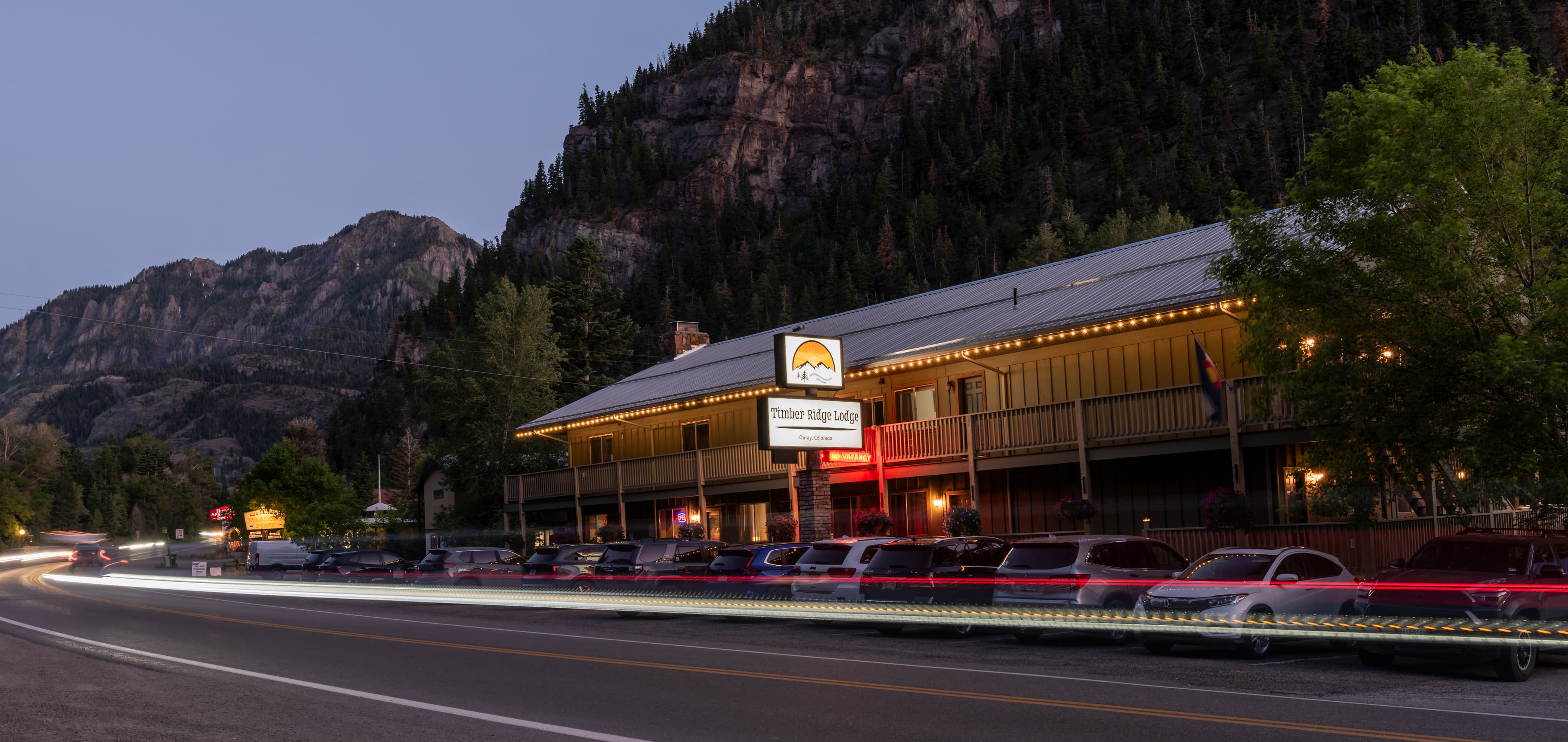 A cozy lodge illuminated at dusk, nestled between mountains along a quiet road.