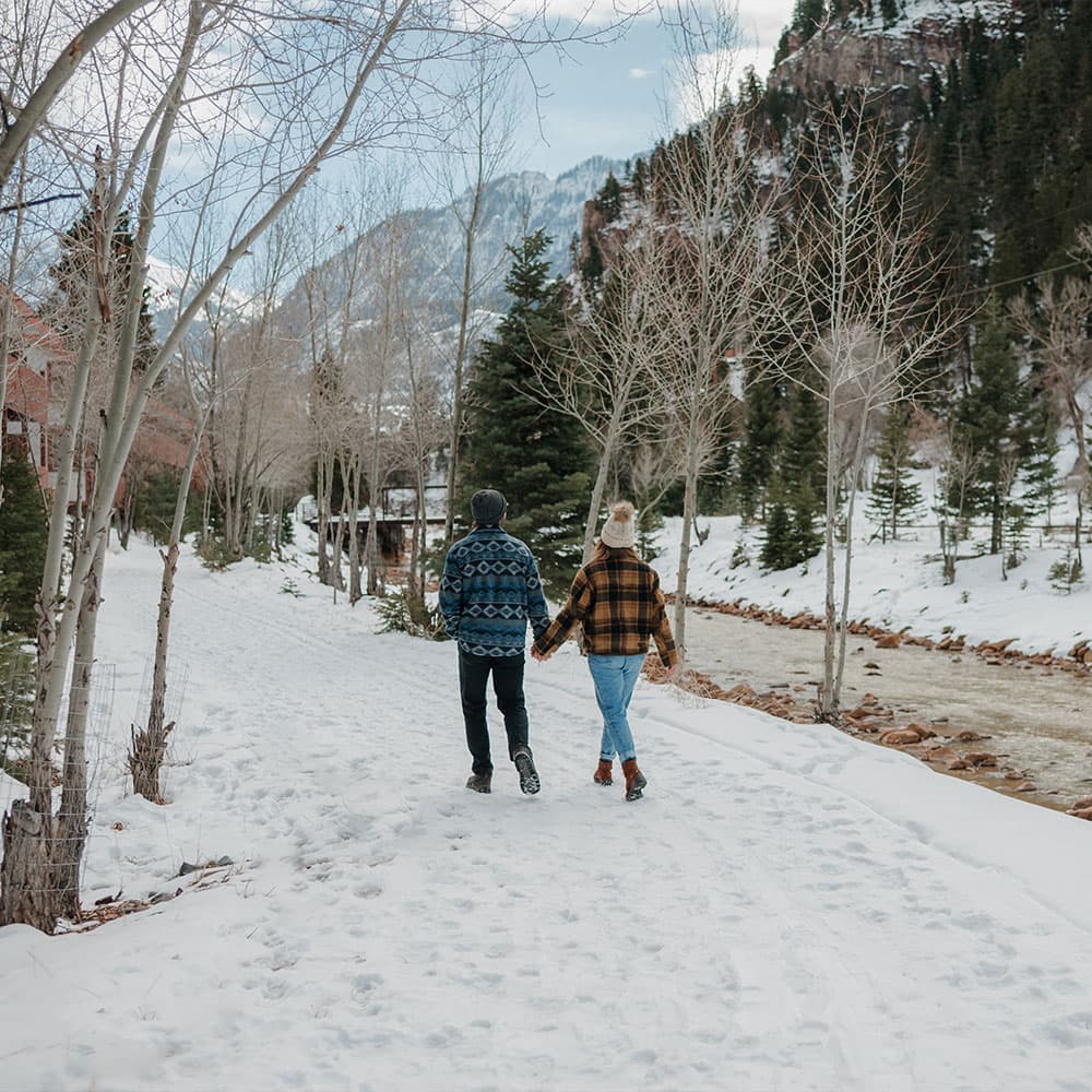 A couple walks hand-in-hand along a snowy path by a river, surrounded by trees and mountains.
