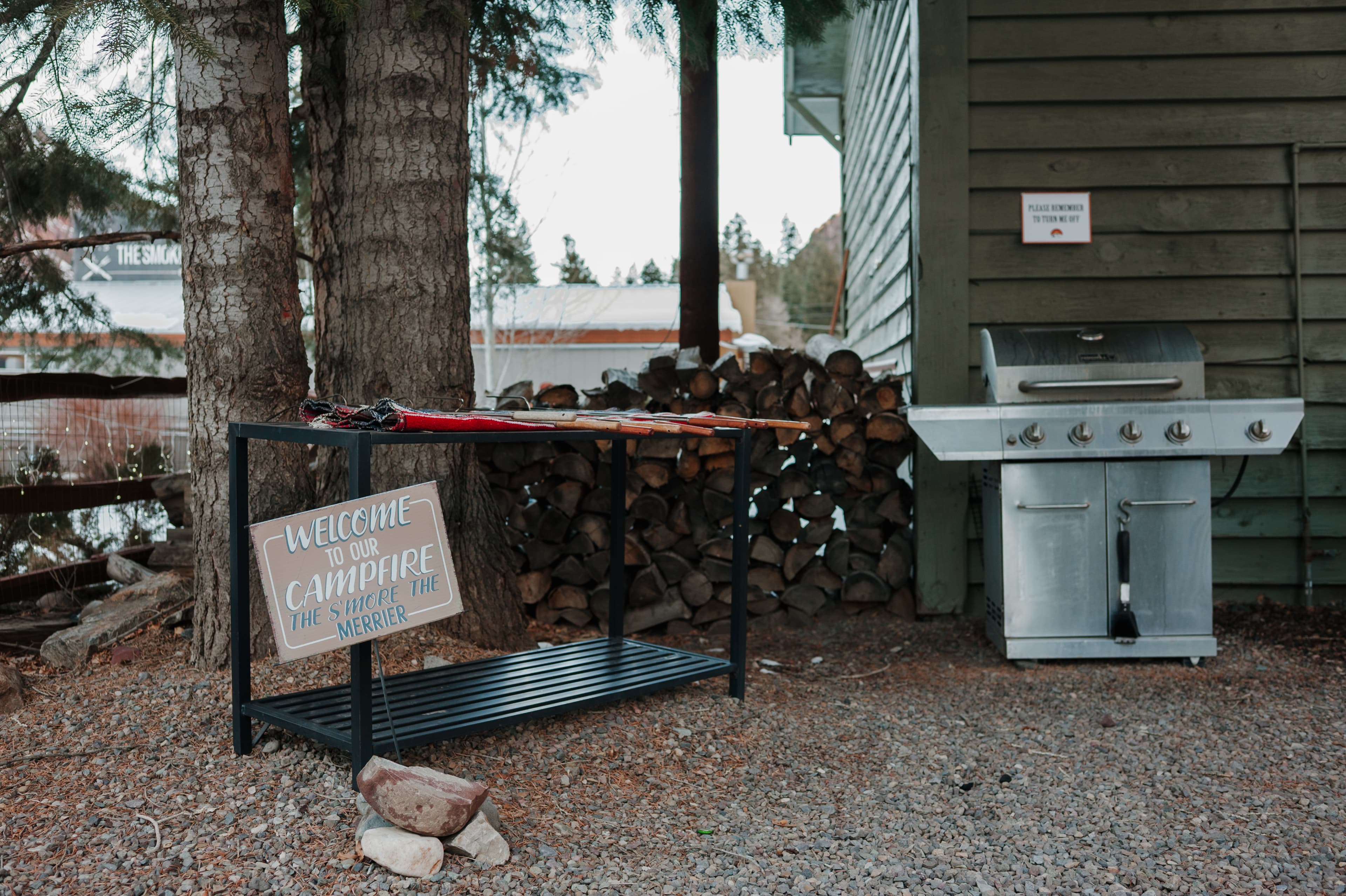 A welcome sign stands beside a grill and stacked firewood against a cabin wall.