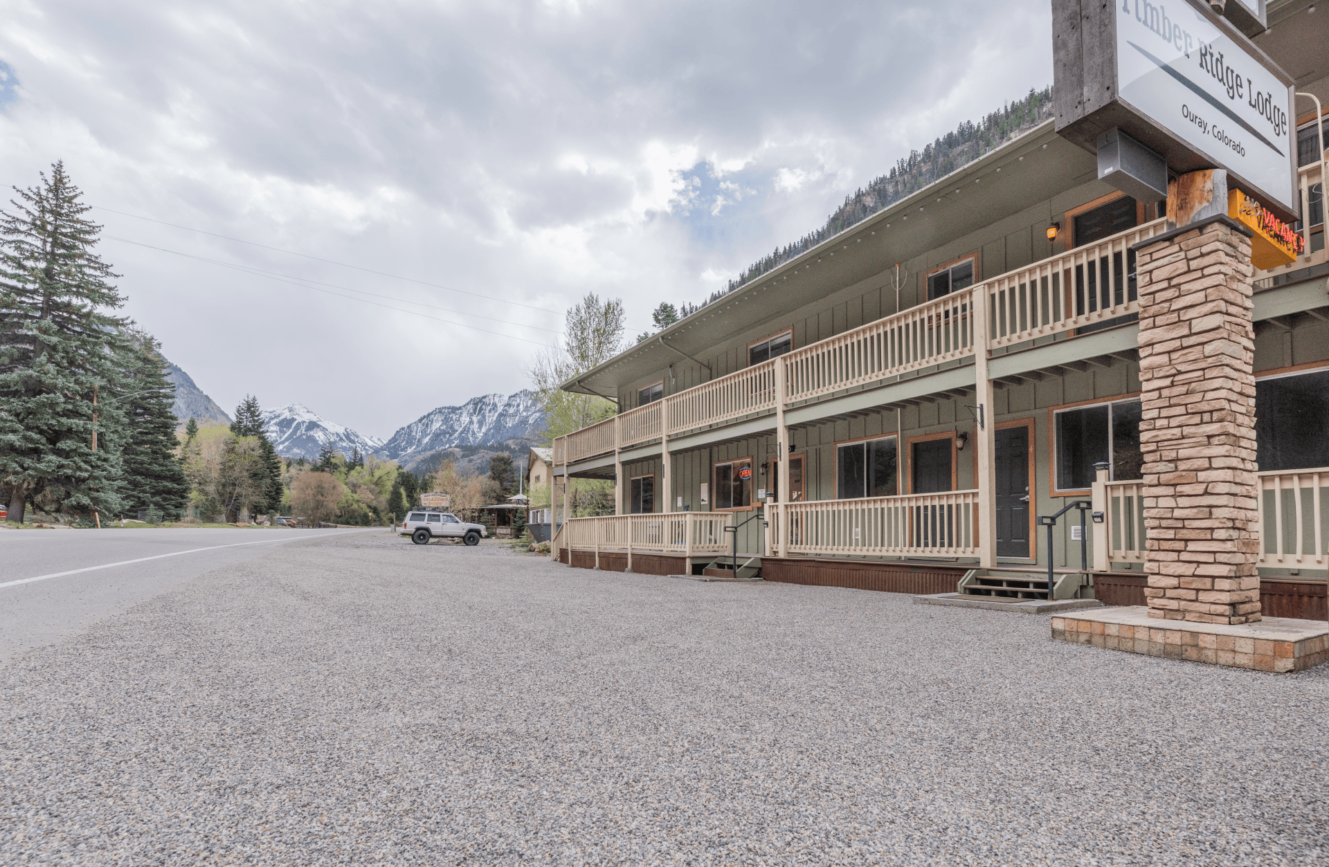A gravel parking area in front of a lodge with a mountainside backdrop under a cloudy sky.