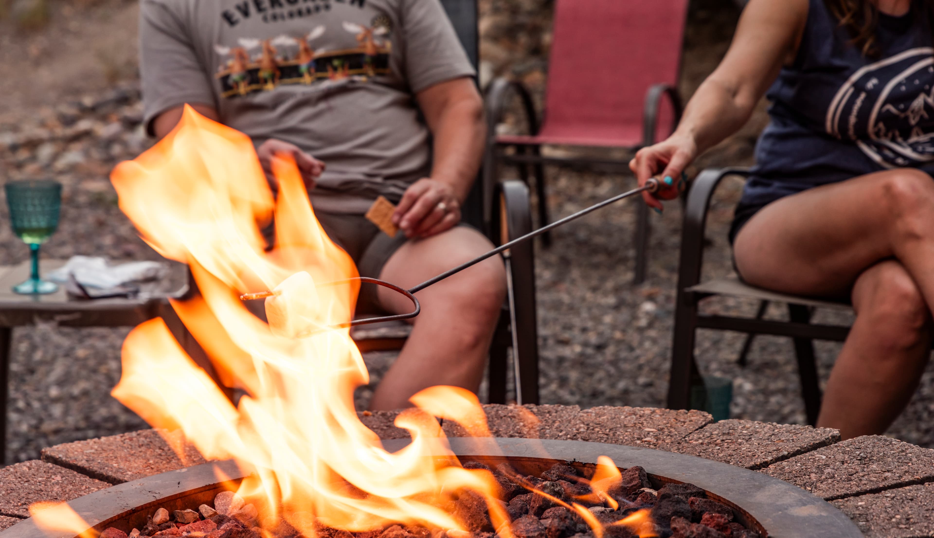 Two people sit by a fire pit, one roasting a marshmallow over the flames.