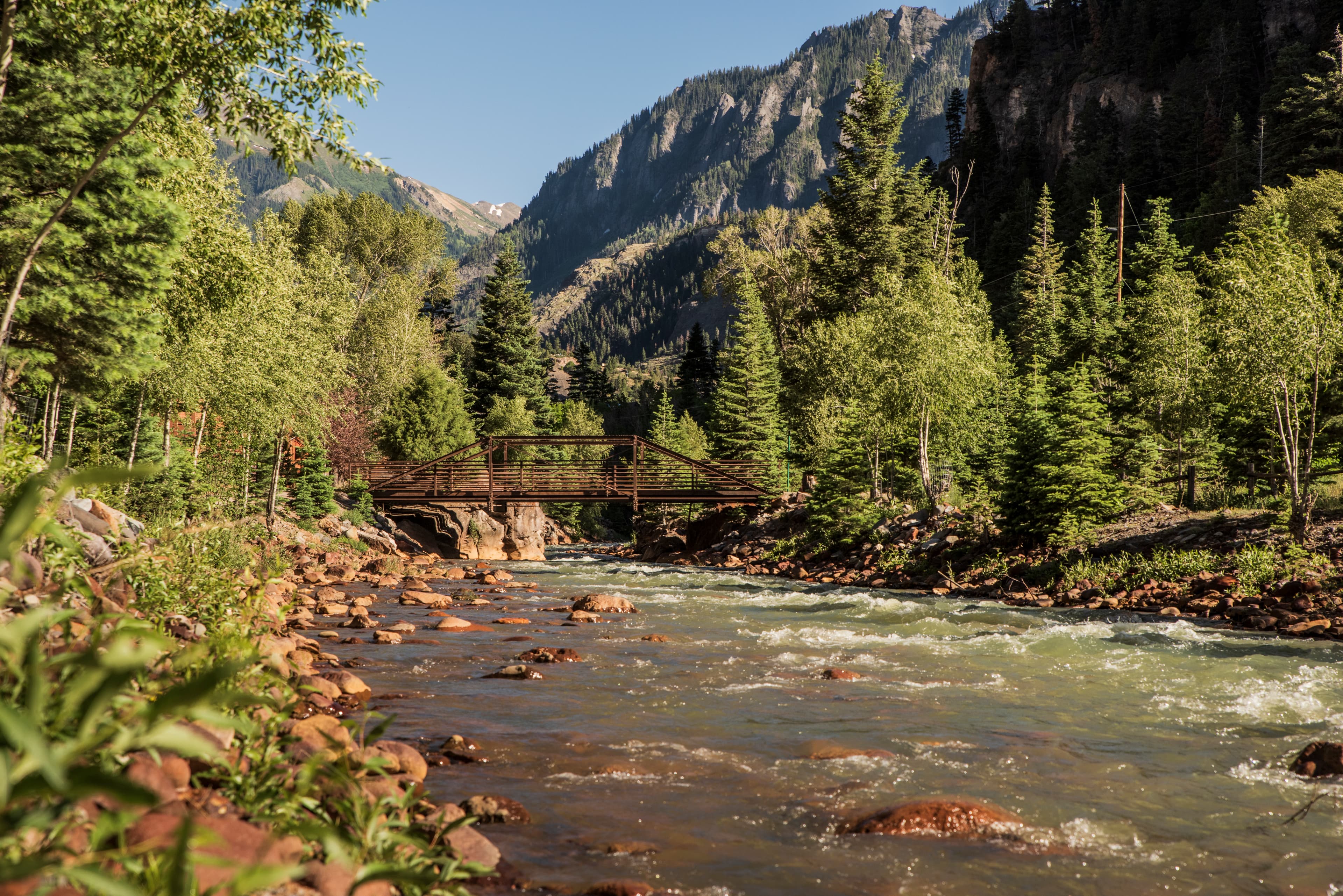 A wooden bridge spans a flowing river surrounded by lush greenery and mountains under a clear blue sky.