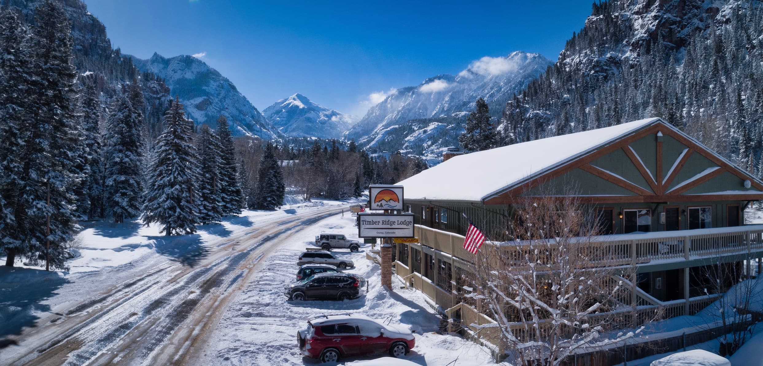 A snow-covered mountain landscape featuring a lodge and parked cars under a clear blue sky.
