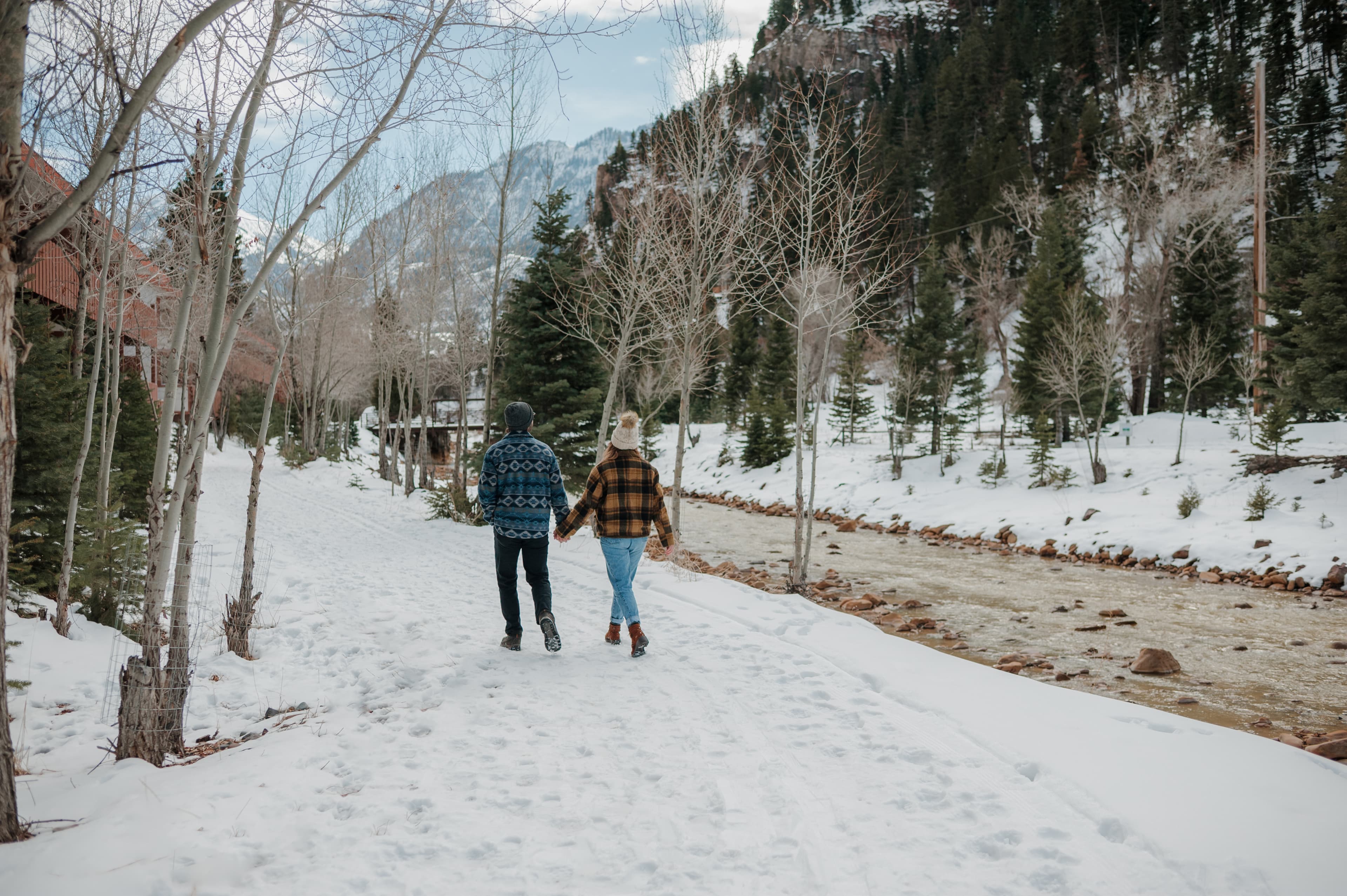 A couple walks hand-in-hand along a snow-covered path beside a flowing river and trees.