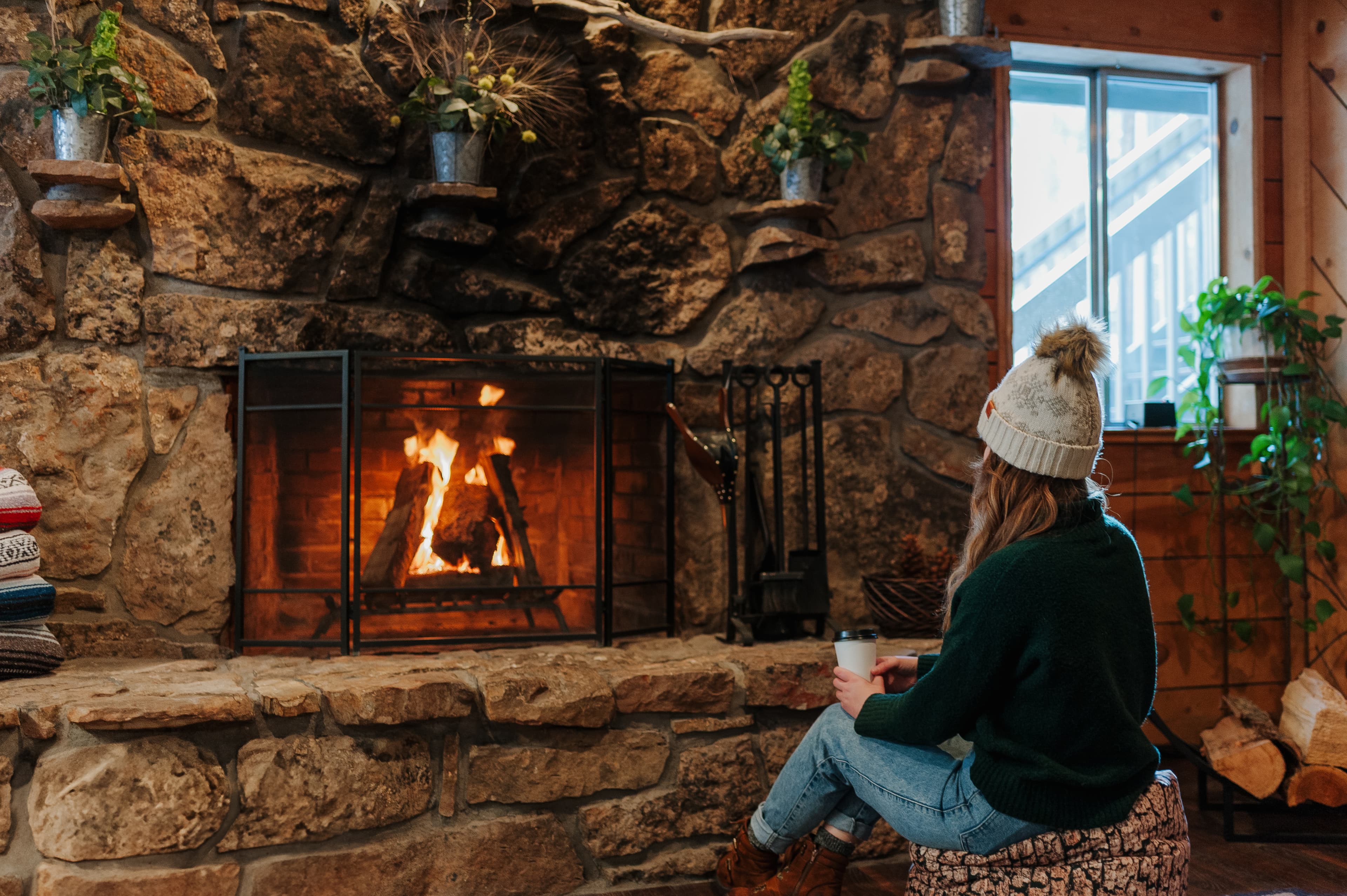 A person sits on a cozy stool by a stone fireplace, sipping a drink while enjoying the warm flames.