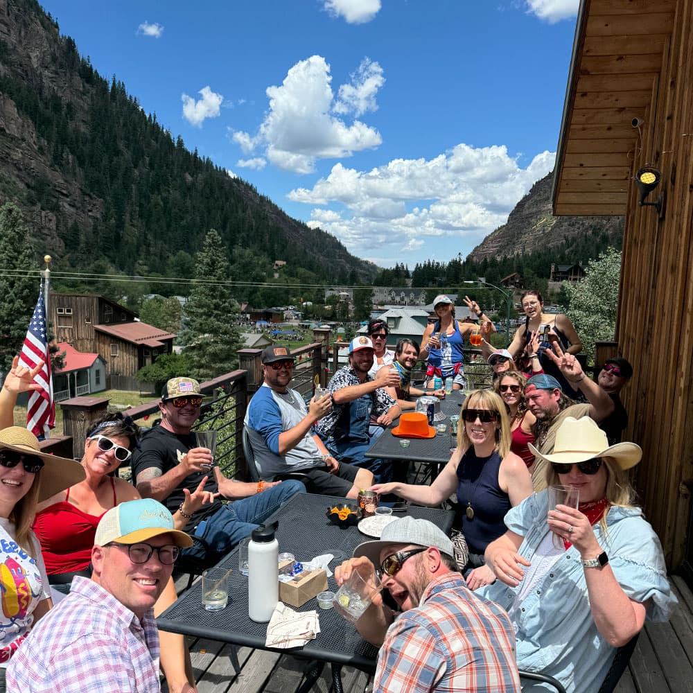 A group of people enjoying drinks and food on a sunny deck overlooking a mountainous landscape.