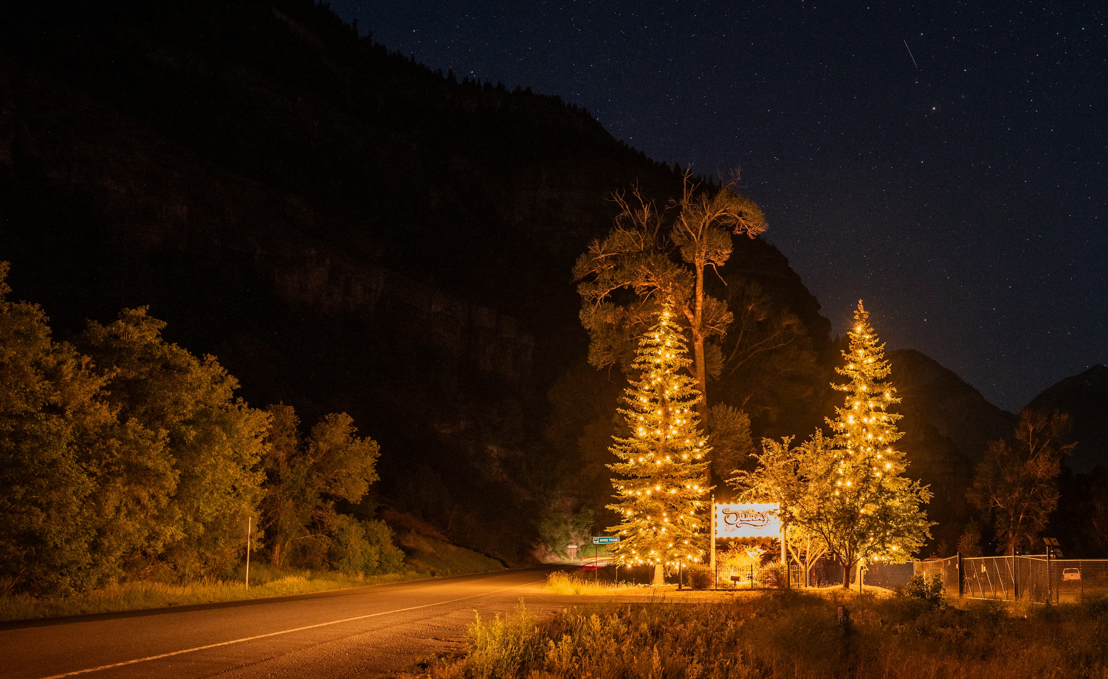A nighttime road scene featuring illuminated trees and a sign against a dark mountainous backdrop.