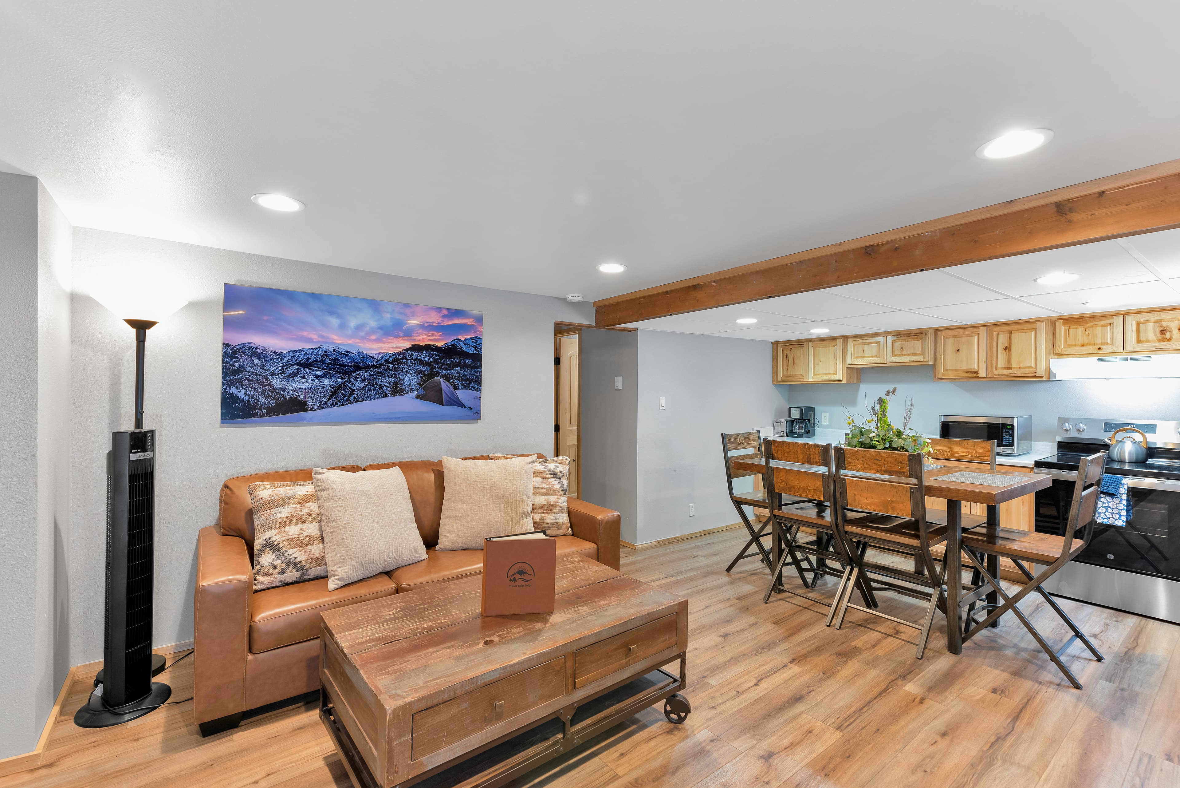 The main living area featuring a tan leather sofa, a reclaimed wood coffee table on wheels, and a tall black floor fan. A large wooden beam separates the living area from a full kitchen and dining table, creating an open-concept mountain lodge feel.