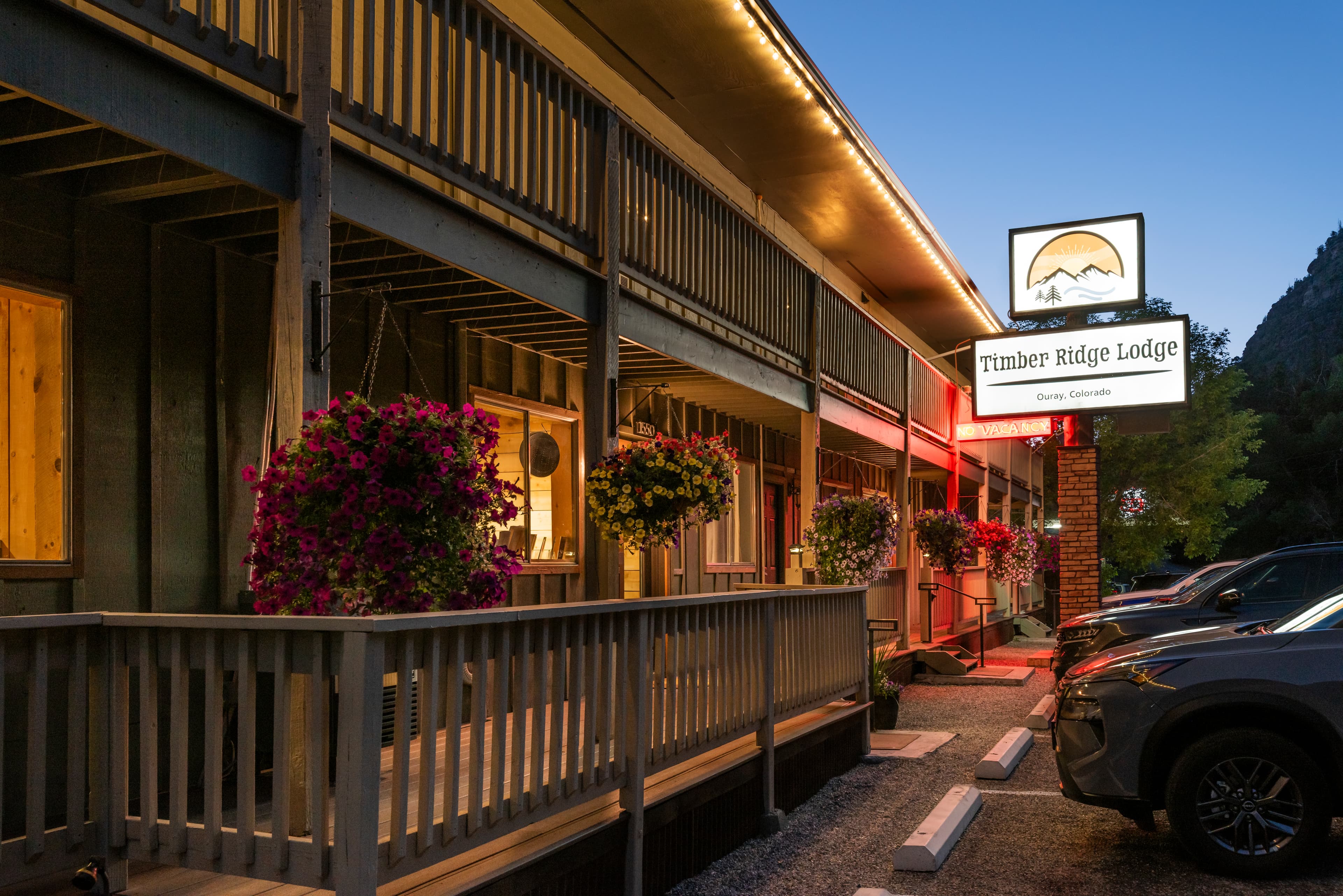 The Timber Ridge Lodge features a wooden exterior with hanging flower baskets, illuminated signage, and parked cars at dusk.