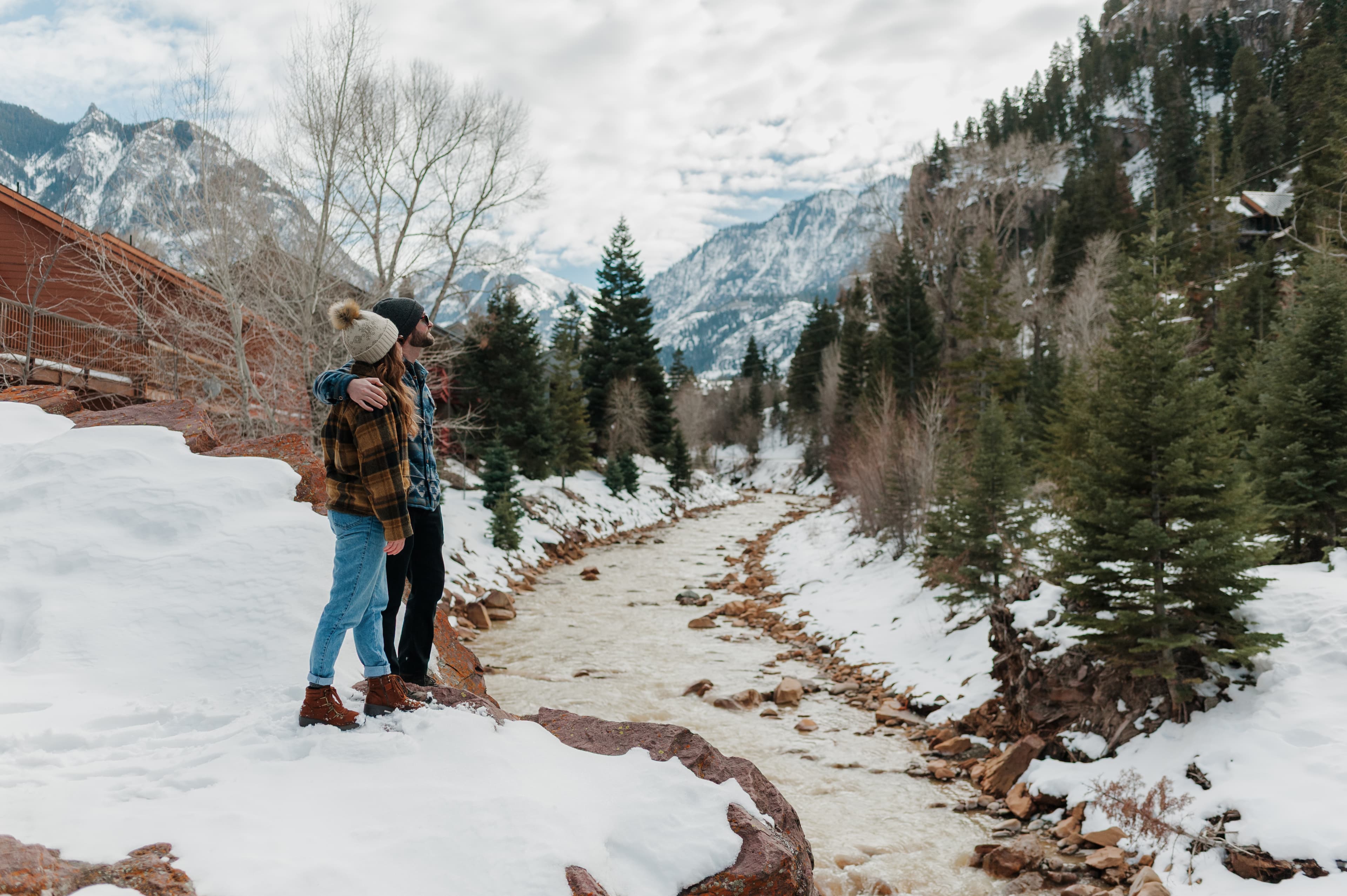 A couple stands on a snow-covered rock by a flowing river, surrounded by mountains and pine trees.