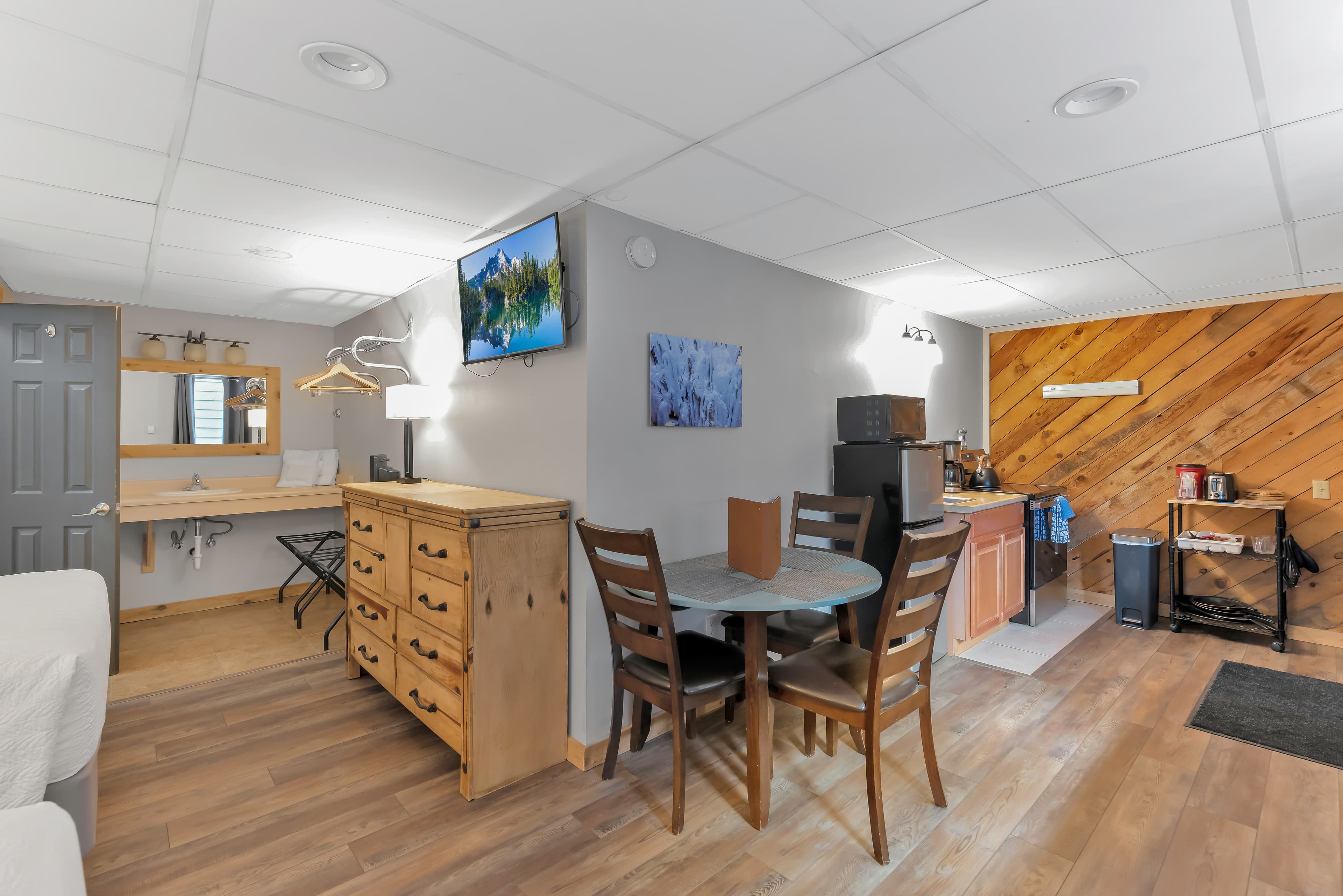 A wide-angle corner view of the Queen Room showing the open-concept layout. The space includes two queen beds, a vanity area with a large mirror, a wooden dresser, a small dining set, and a kitchenette equipped with a mini-fridge, microwave, and stove against a diagonal wood-plank wall.