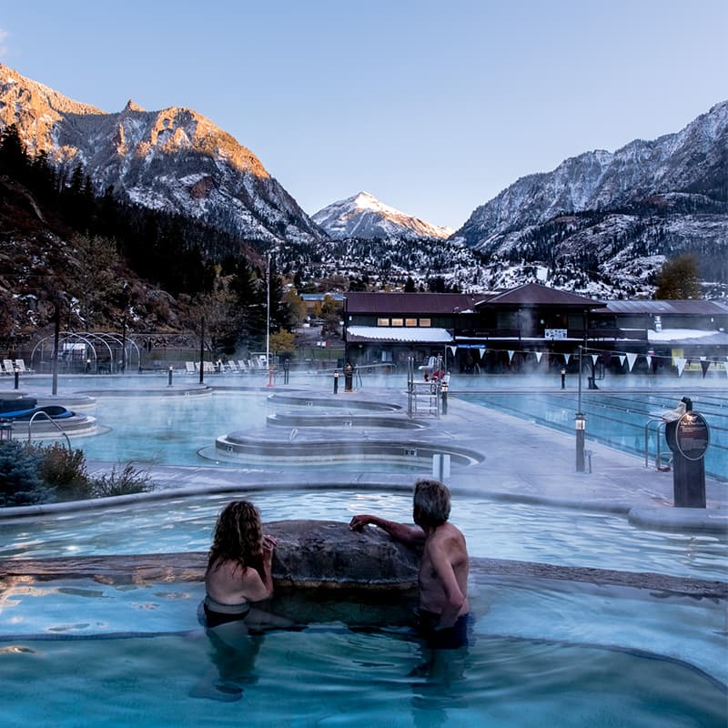Two people relax in a hot spring with mountain scenery and steam rising in the background.