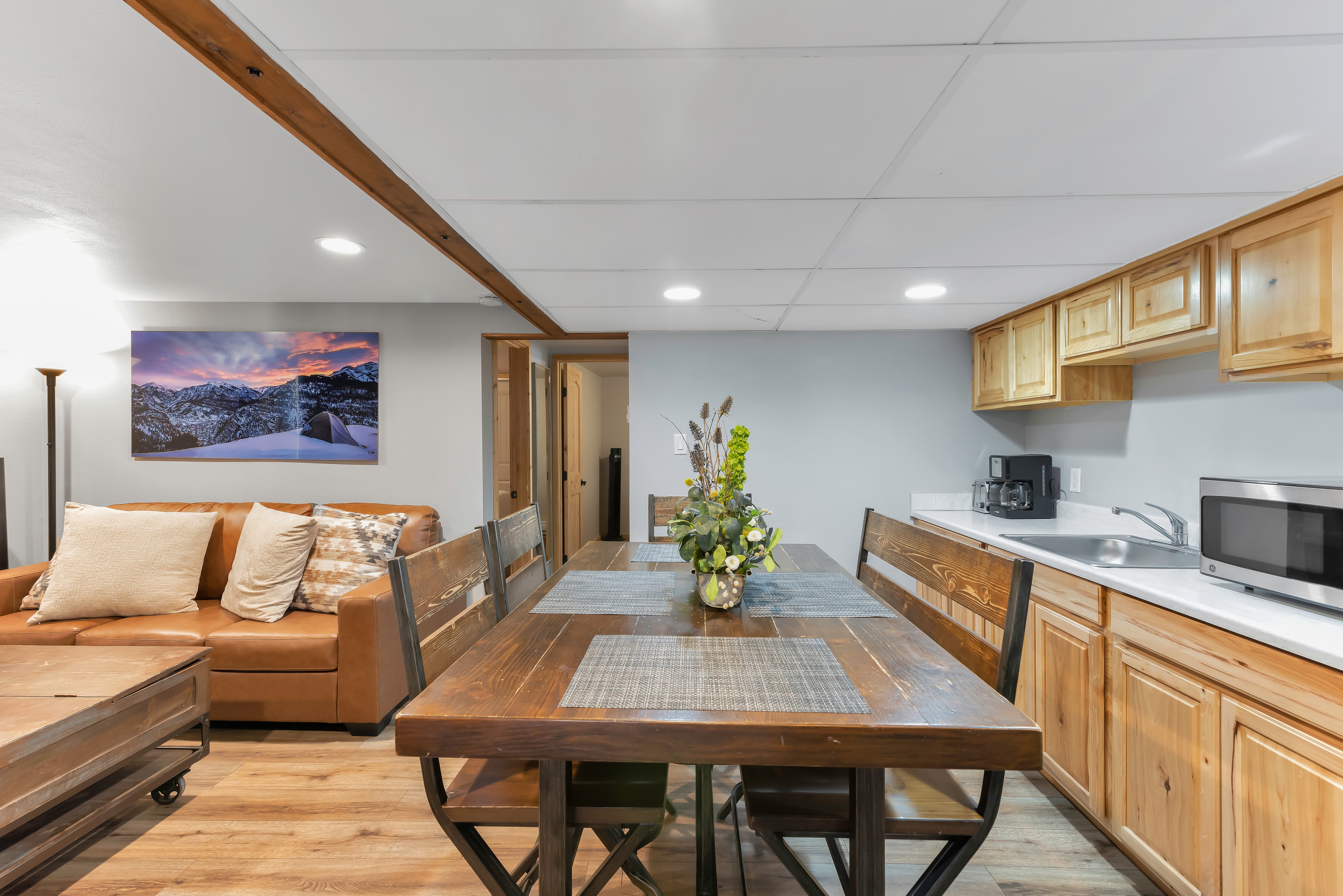 A side view of the apartment living space showing a long, rustic wooden dining table with seating. To the left, a leather sofa sits under a sunset mountain landscape photo. To the right is a kitchenette with light wood cabinets, a white countertop, a sink, and a microwave.