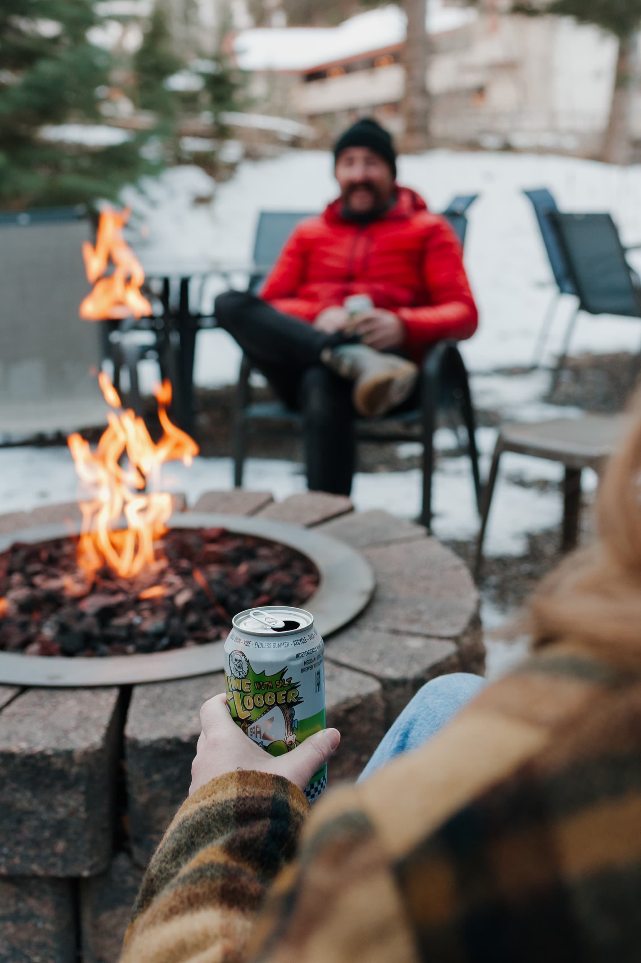 A person in a plaid jacket holds a can while sitting by a fire pit, with a friend in a red jacket visible in the background.