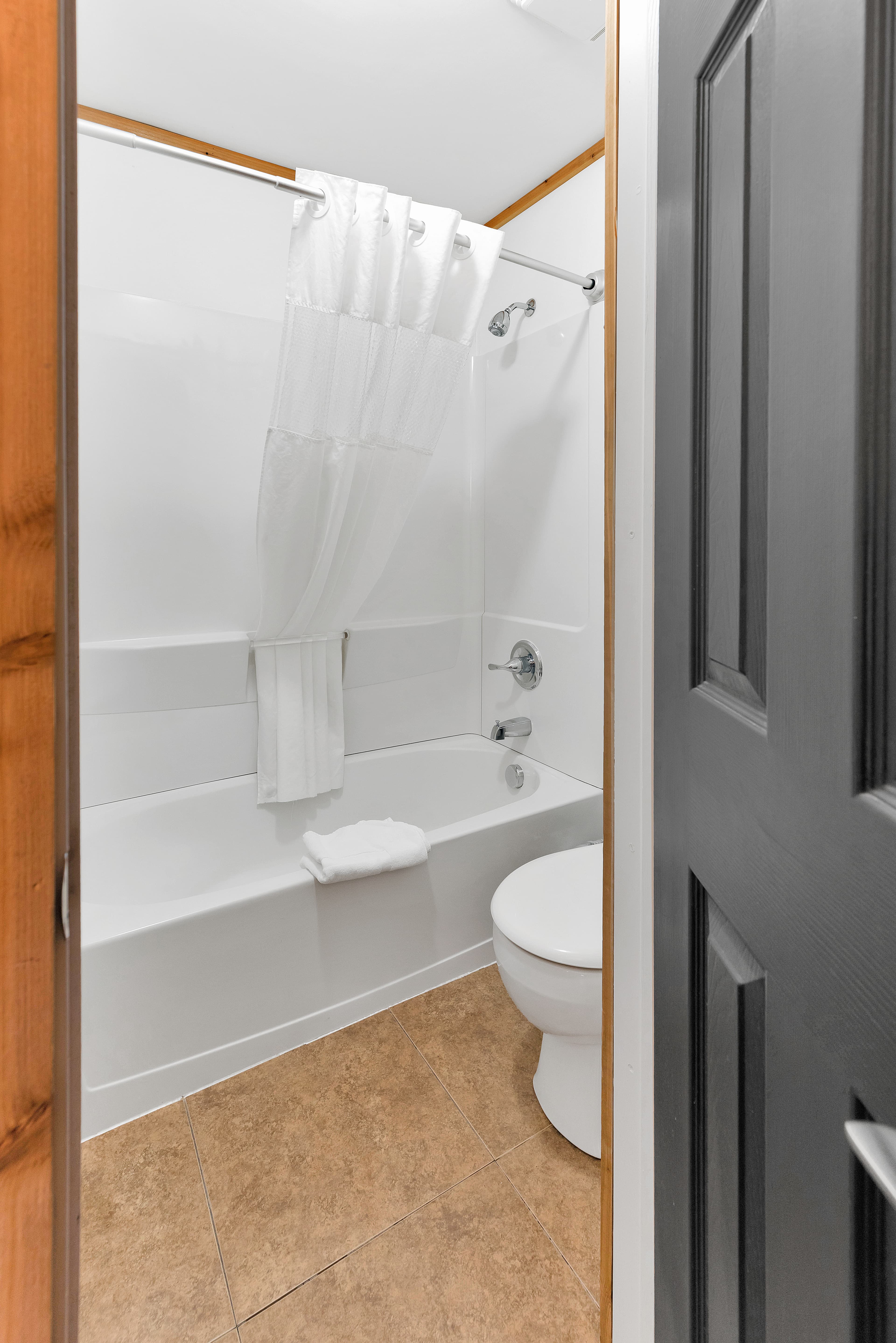 A view through a dark grey door into the bathroom, showing a white bathtub and shower surround with a white curtain. The room features tan tile flooring and a white toilet, with natural wood trim around the door frame.