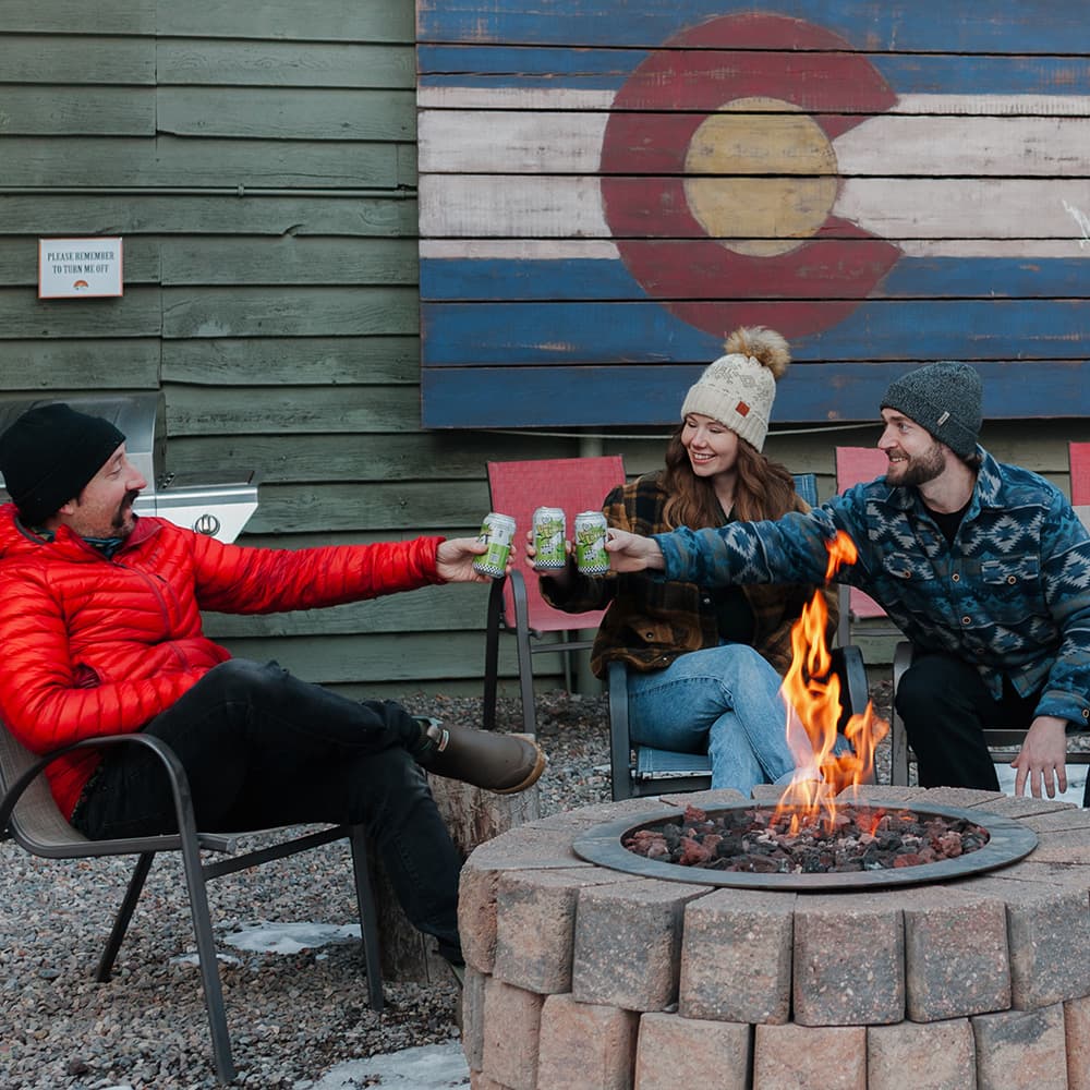 Three friends toast with drinks around a fire pit in front of a Colorado-themed backdrop.