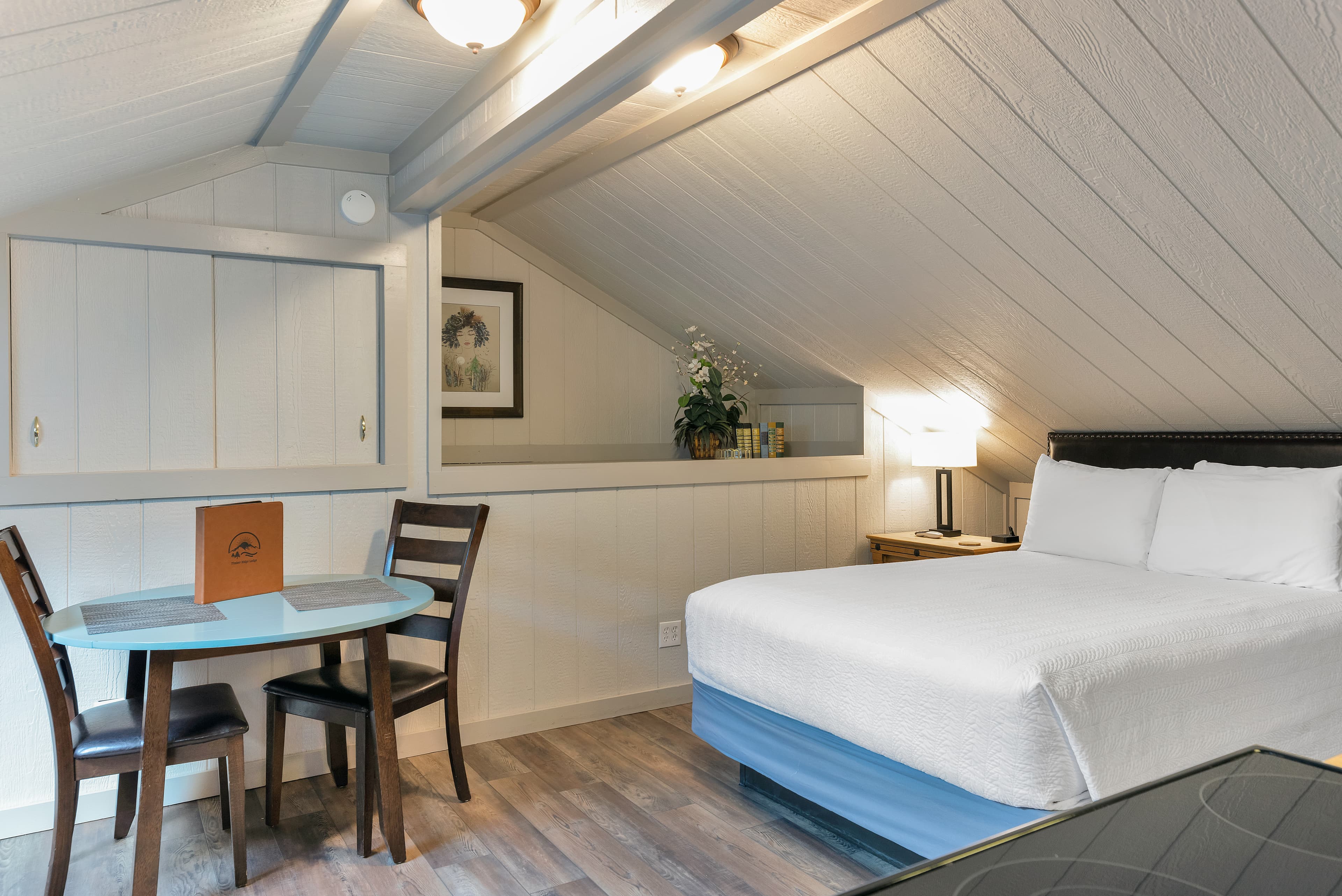 A view of the sleeping and dining areas in a rustic-modern loft. The space includes a queen-sized bed with white linens, a small bistro dining set with two chairs, and light-colored wood-paneled walls that create a bright, airy mountain cabin atmosphere.