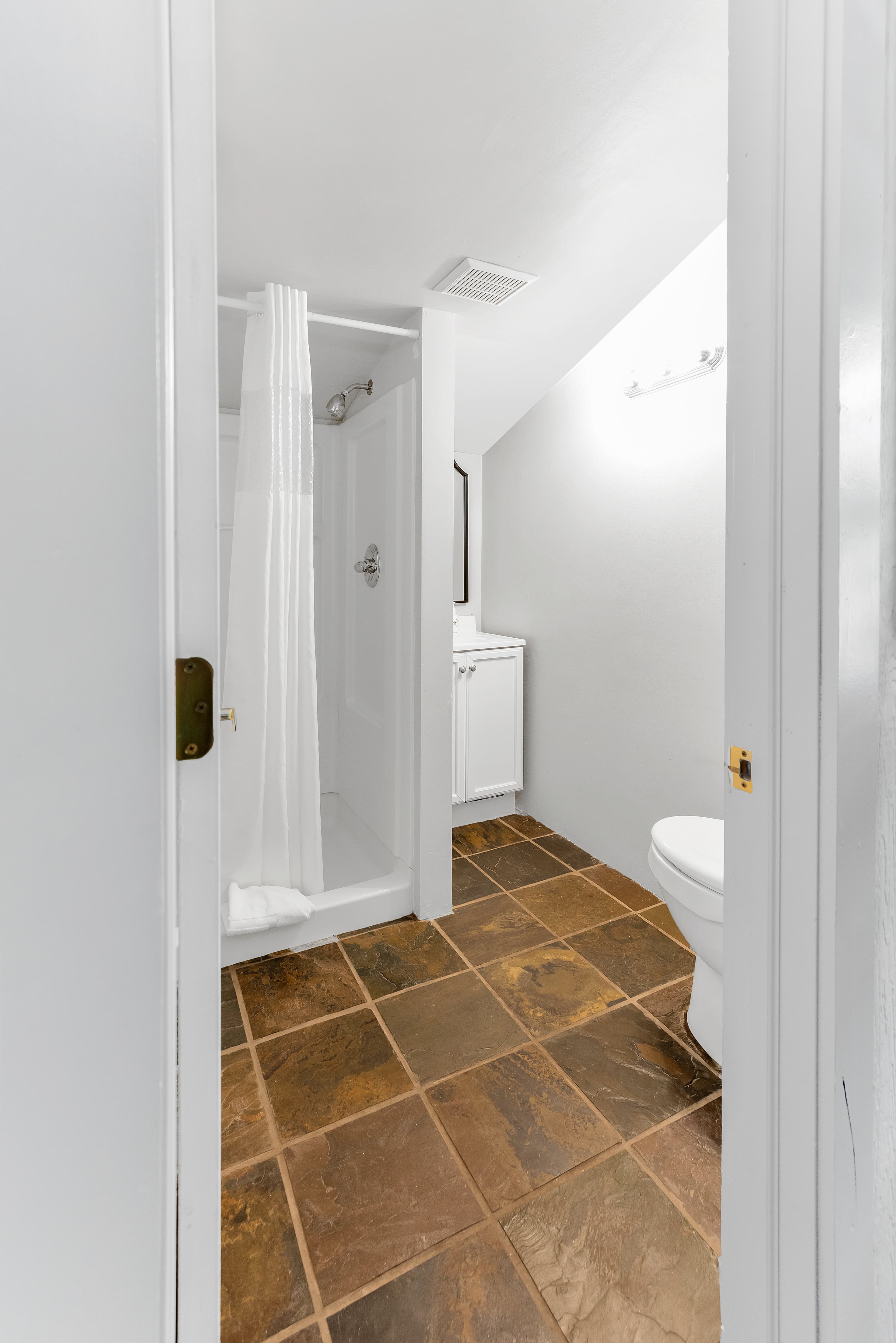 A bright, efficient bathroom in the loft suite featuring natural slate tile flooring. The space includes a walk-in shower with a white curtain, a white vanity with a mirror, and a toilet, all set against clean white walls under a sloped ceiling.