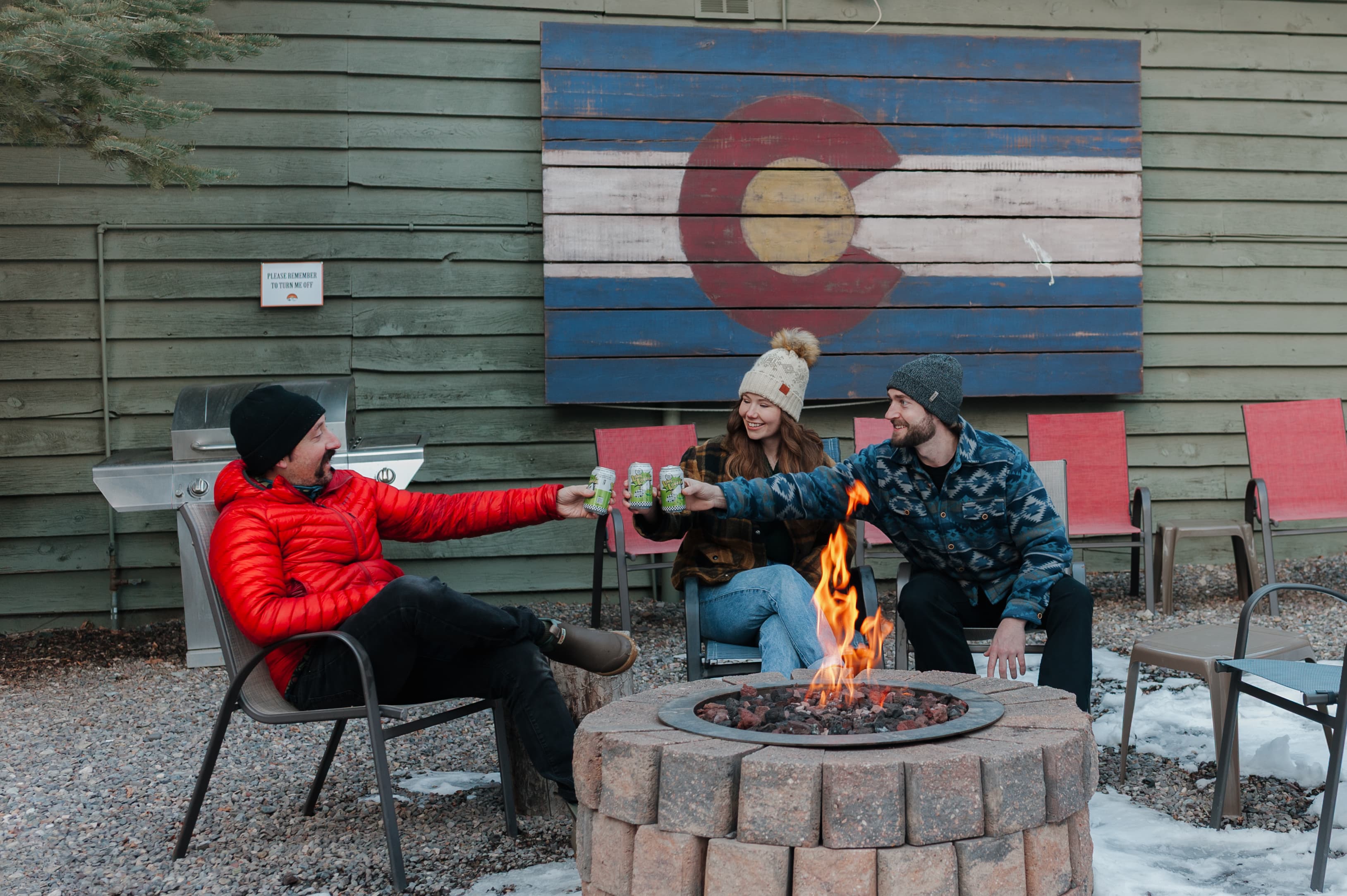 Three friends toast with drinks around a fire pit in front of a Colorado-themed backdrop.