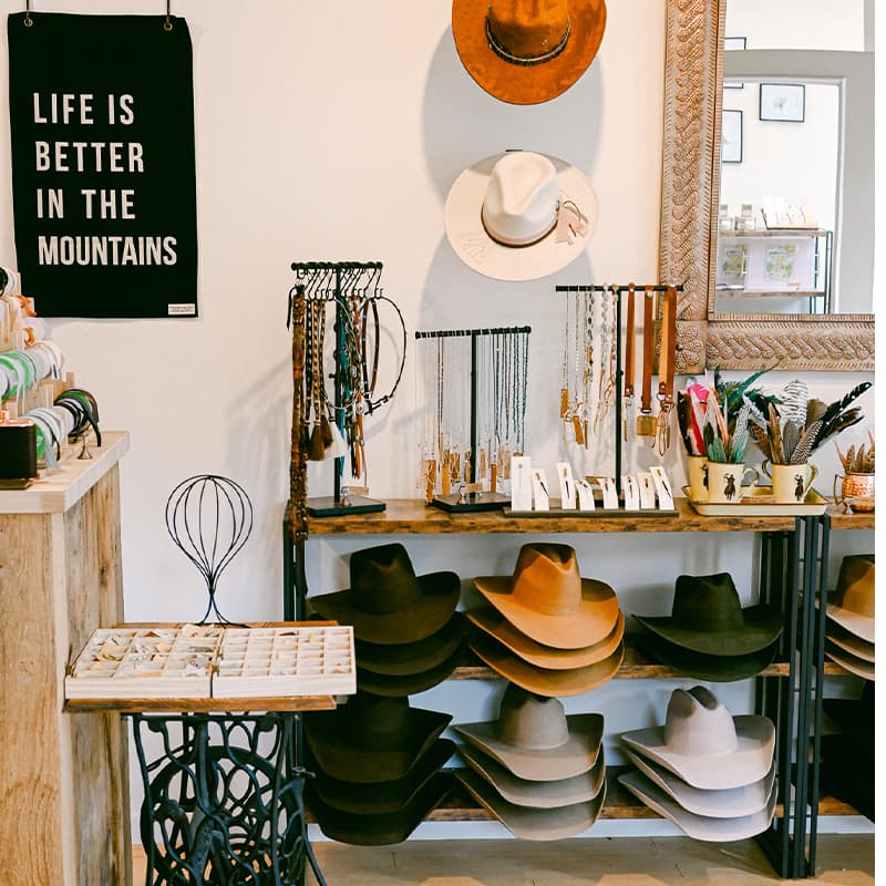 A cozy shop featuring hats, jewelry, and a wall sign that reads "LIFE IS BETTER IN THE MOUNTAINS."