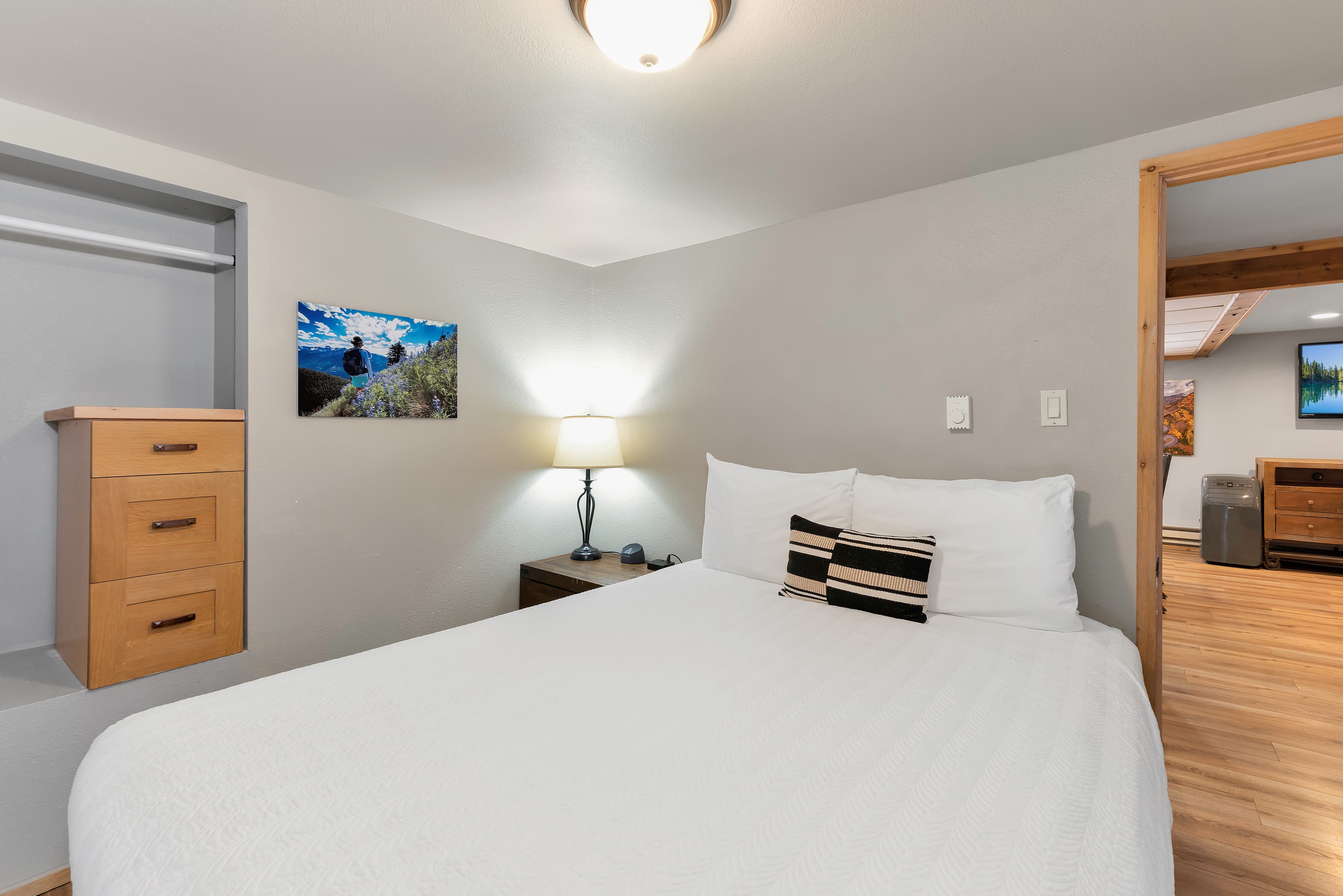 The first bedroom featuring a queen bed with white linens and a striped accent pillow. A large canvas print of a starry night sky over a mountain valley hangs above the headboard, flanked by a dark wood dresser and white sliding closet doors.